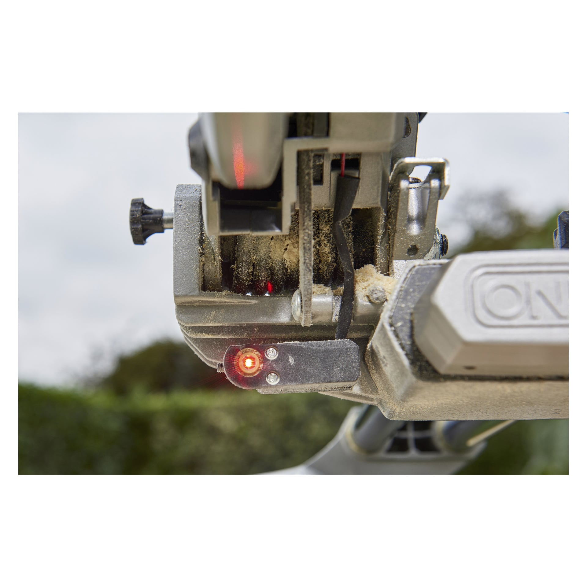 Lähikuva Ryobi One+ R18MS216-0 -paristokäyttöisestä jiirisahasta käytössä, jossa näkyy terä, ON-kytkin, hohtava punainen turvavalo ja kertynyt sahapuru. Pehmeästi sumea tausta vihreällä lehvästöllä viittaa siihen, että kohtaus on ulkona.