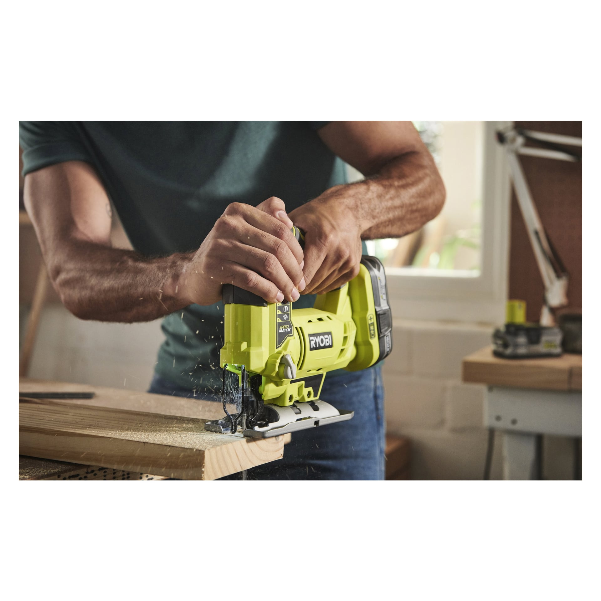 En person i mörk skjorta använder en grön Ryobi One+ Jigsaw R18JS-120S5 för att såga trä på en arbetsbänk, med träspån och sågspån utspridda runtomkring. Ytterligare verktyg och ett fönster syns i bakgrunden.