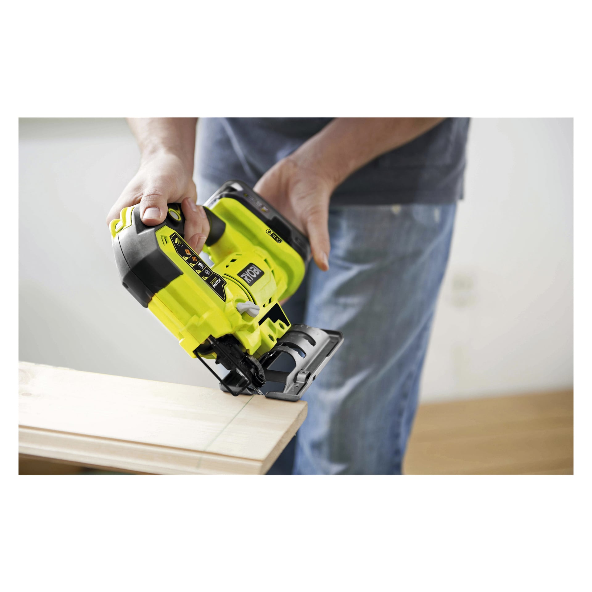 En person kör en gul och svart trådlös Ryobi One+ Jigsaw R18JS-120S5 för att skära en rak linje genom en bit ljust trä. Endast personens händer och överkropp syns på bilden.