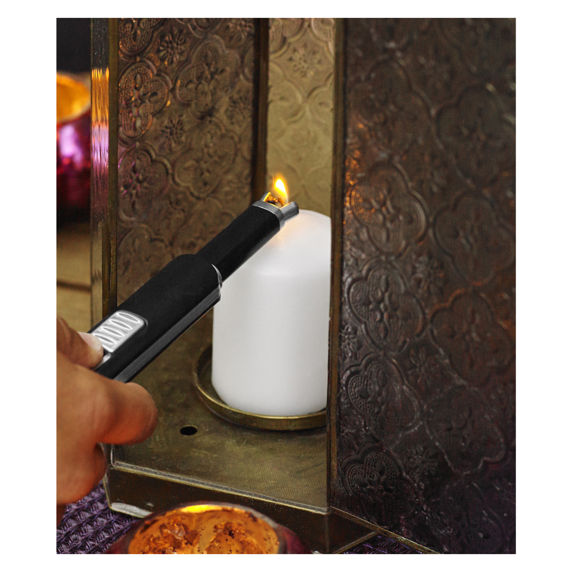 En hånd betjener en svart Menuett USB-lighter for å tenne et hvitt søylelys plassert inne i en dekorativ metalllykt, med ekstra lys og ulike dekorative detaljer i nærheten.