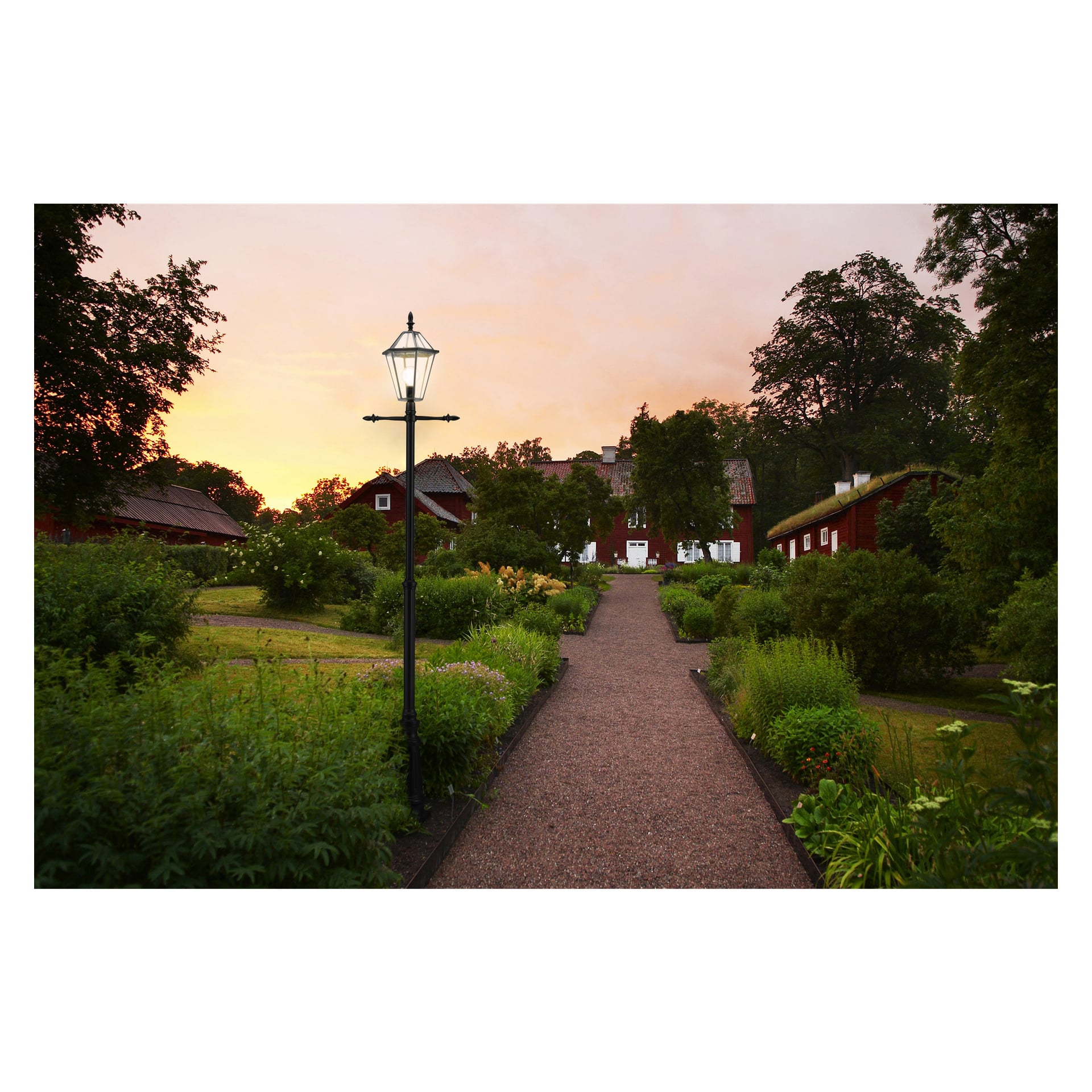 En slingrande grusstig skär genom livfullt gröna trädgårdar och leder till röda träbyggnader som varmt upplysts av den nedgående solen. En vintage-inspirerad Anslut-lyktstolpe kantar gångvägen och kastar ett milt ljus under lövträd som omsluter det fridfulla landskapet.