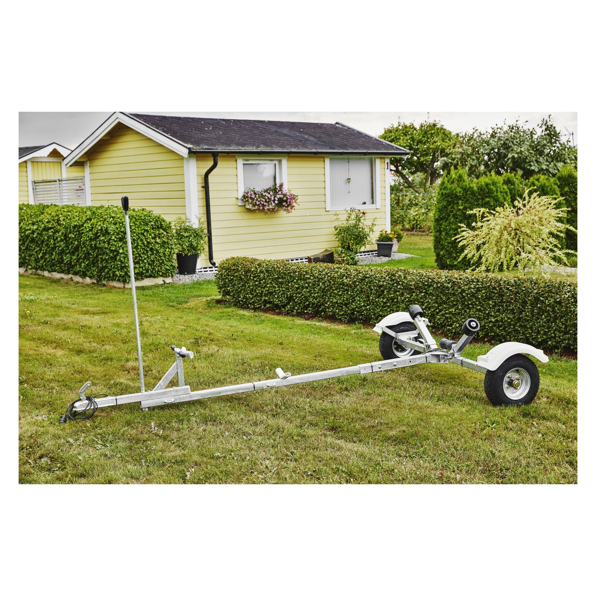 En Kayoba Marine Dinghy-trailer med två hjul står på gräsmattan framför ett gult hus med vita lister, färgglada blommor i en fönsterlåda och frodiga gröna häckar och träd som omger fastigheten.