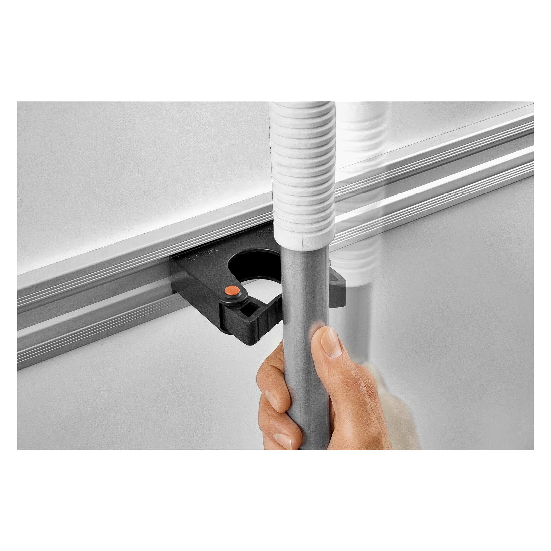 Ręka wkłada drążek z białą rączką do uchwytu narzędziowego Toolflex (2 sztuki), który składa się z czarnych uchwytów ściennych z pomarańczowymi przyciskami, przymocowanych do srebrnej szyny.