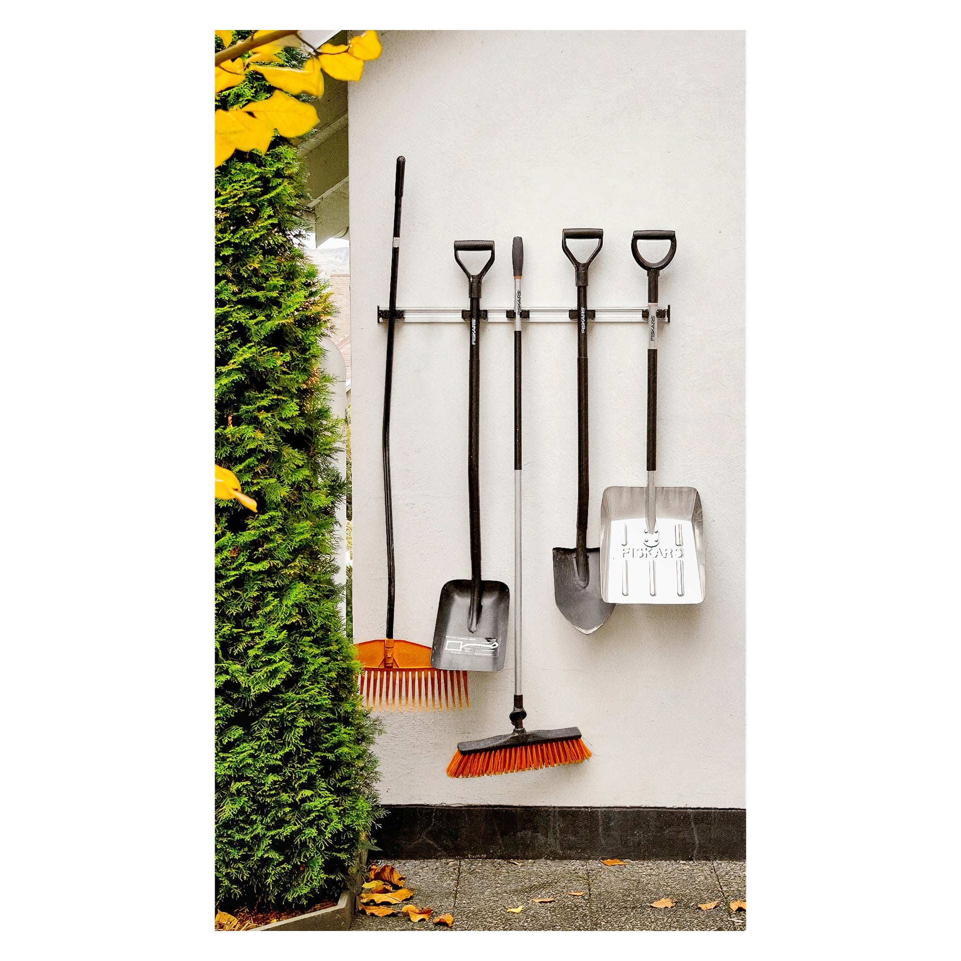 Pięć narzędzi ogrodniczych — dwie miotły, dwie łopaty i łopata do śnieżu — jest uporządkowanych na Toolflex Tool Holder 2 Pack, który jest zamontowany na ścianie obok wysokiego zielonego krzewu. Teren to zewnętrzny taras wyłożony kafelkami.
