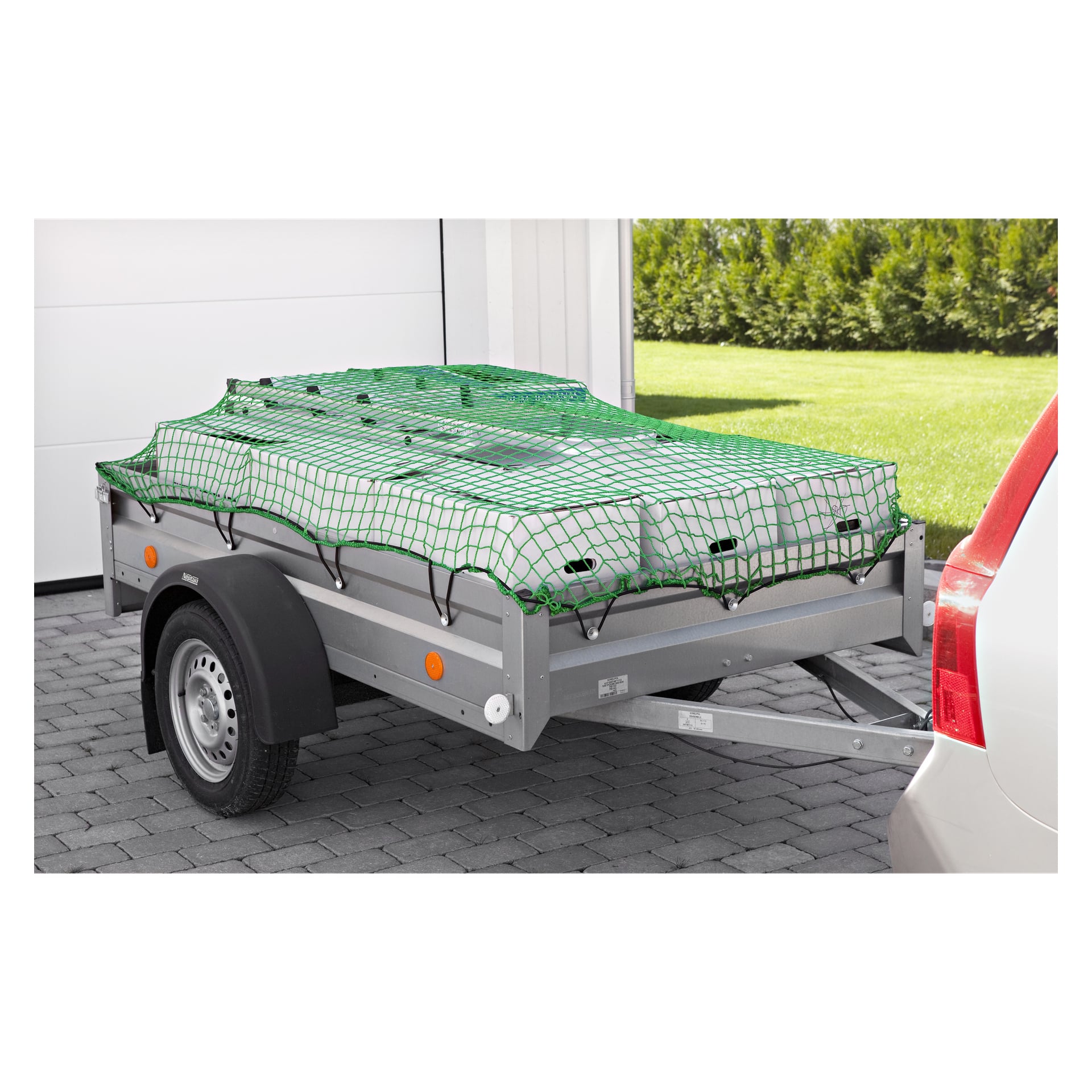 Ett Hamron Load-nät håller lasten på plats på en liten släpvagn kopplad till en vit bil, som står parkerad på en uppfart bredvid grönt gräs och en vit byggnad.