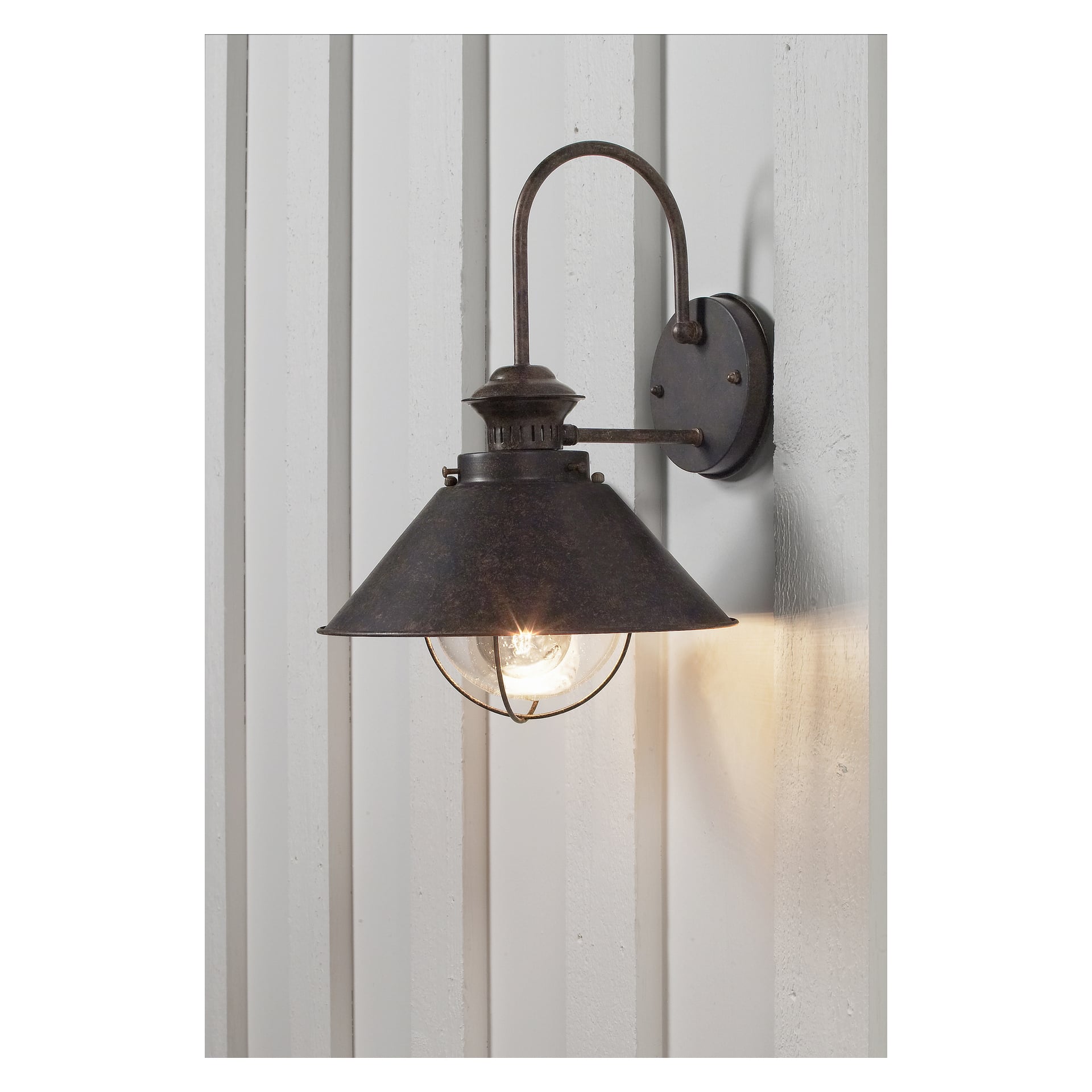 En rustik metallvägglampa av Anslut, med en kupolformad skärm och ett klart glaslampskydd, är monterad på en vertikalt panelad yta. Lampan är tänd och avger ett mjukt sken.