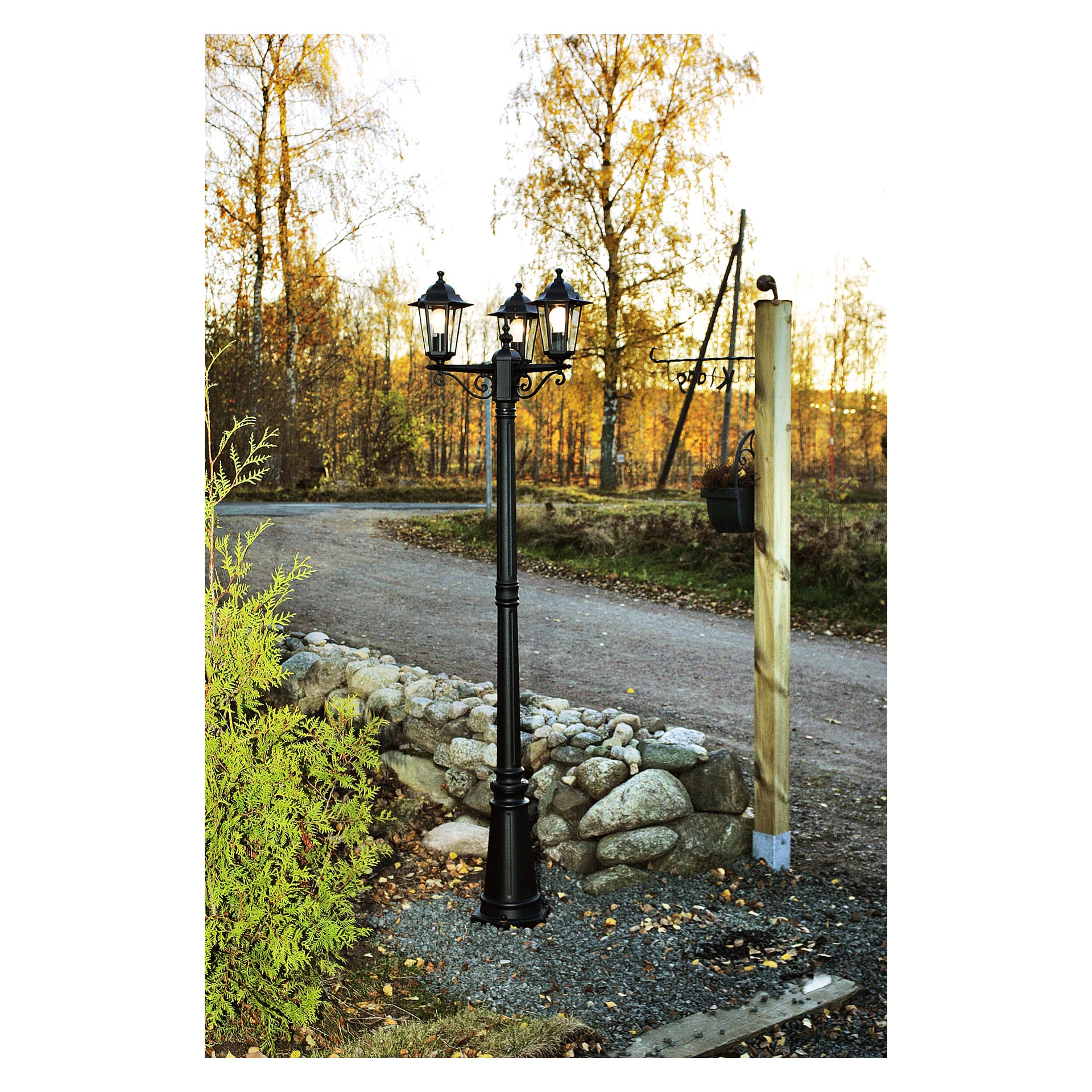 En svart, vintage-inspirert Anslut-lyktestolpe med tre lykter står på en grusvei omgitt av steiner, med høstfargede trær og en trestolpe i bakgrunnen.