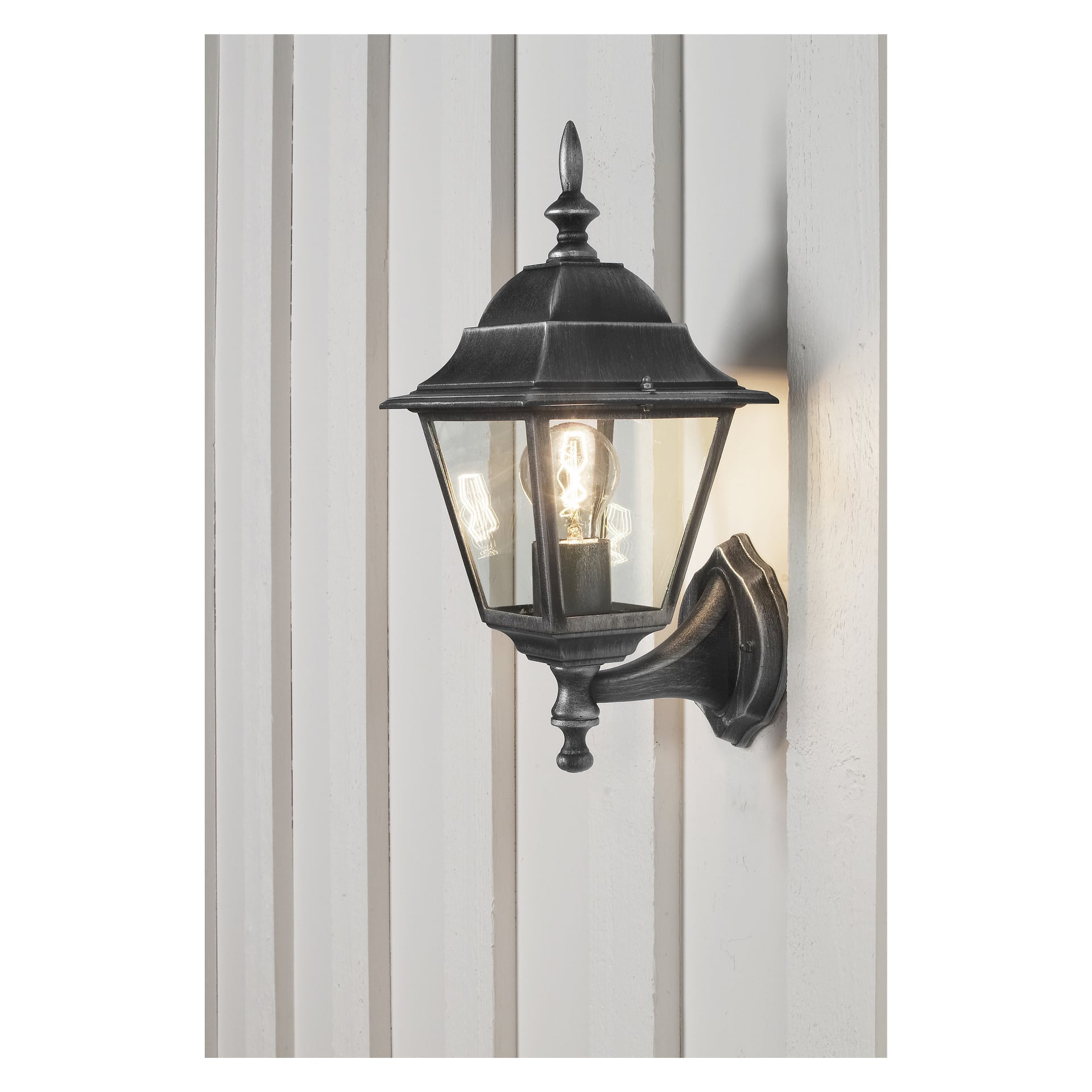 En Anslut-vegglampe med svart metall- og glasskonstruksjon er festet til en vertikalt panelt vegg. Den glødende lyspæren lyser mykt opp området rundt.