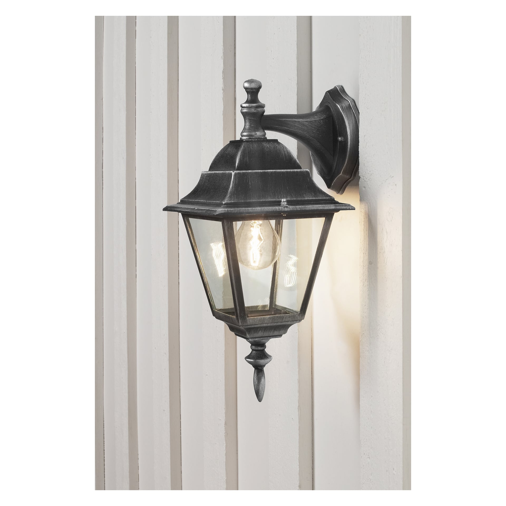 Ansluts vägglampa har en svart metallram och ett klart glasskydd, fäst på en panelad vägg. Lampan är tänd och avslöjar glödlampan inuti.