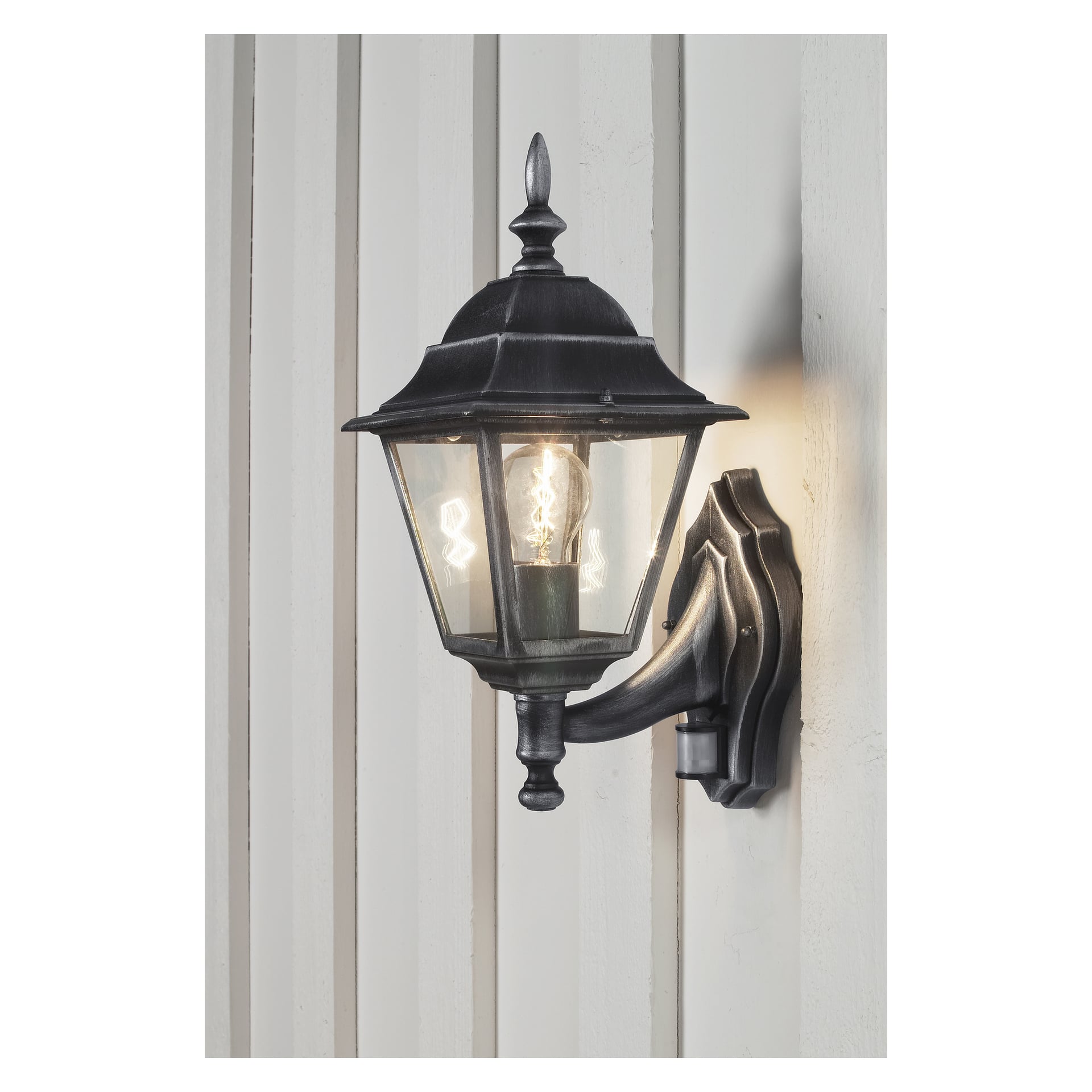 En svart metallvägglampa från Anslut, med rörelsedetektor och klara glaspaneler, är fäst vid en ljus, vertikalt panelad yttervägg. Ljuset inuti lampan är upplyst.