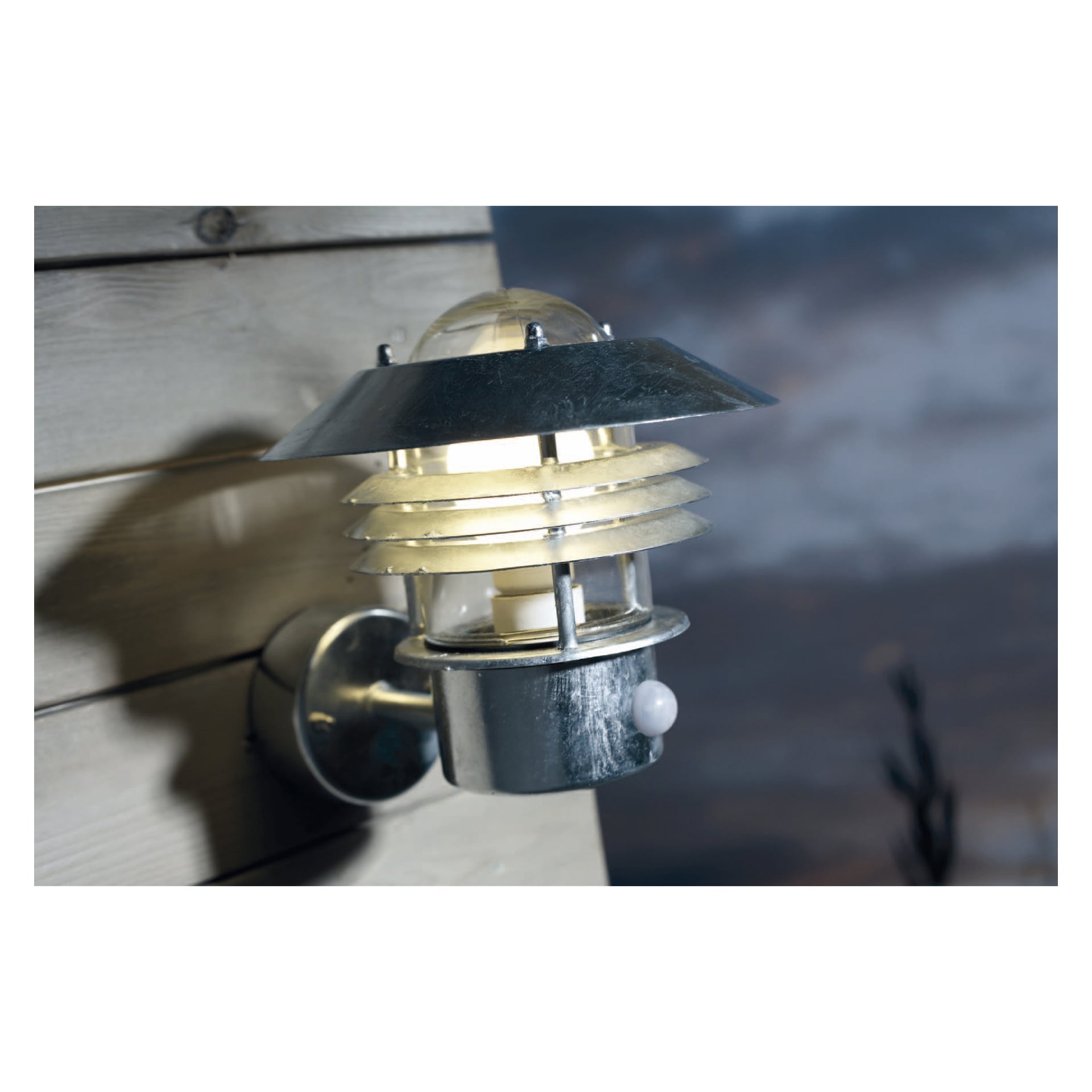 En Nordlux Vejers vägglampa med rörelsedetektor är installerad på träpanel. Armaturen har en rund, trappad design, synliga bultar och en klar kupol högst upp. Lampan är tänd, med en mjukt suddig bakgrund.