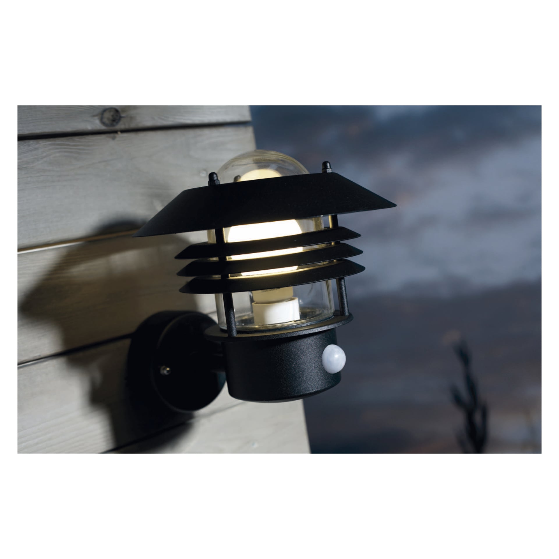En modern svart Nordlux Vejers vägglampa med rörelsedetektor, designad med horisontella metalllistor som omsluter ett glashölje, är installerad på träpanel och lyser varmt. Den mjukt suddiga bakgrunden har en mörk himmel.