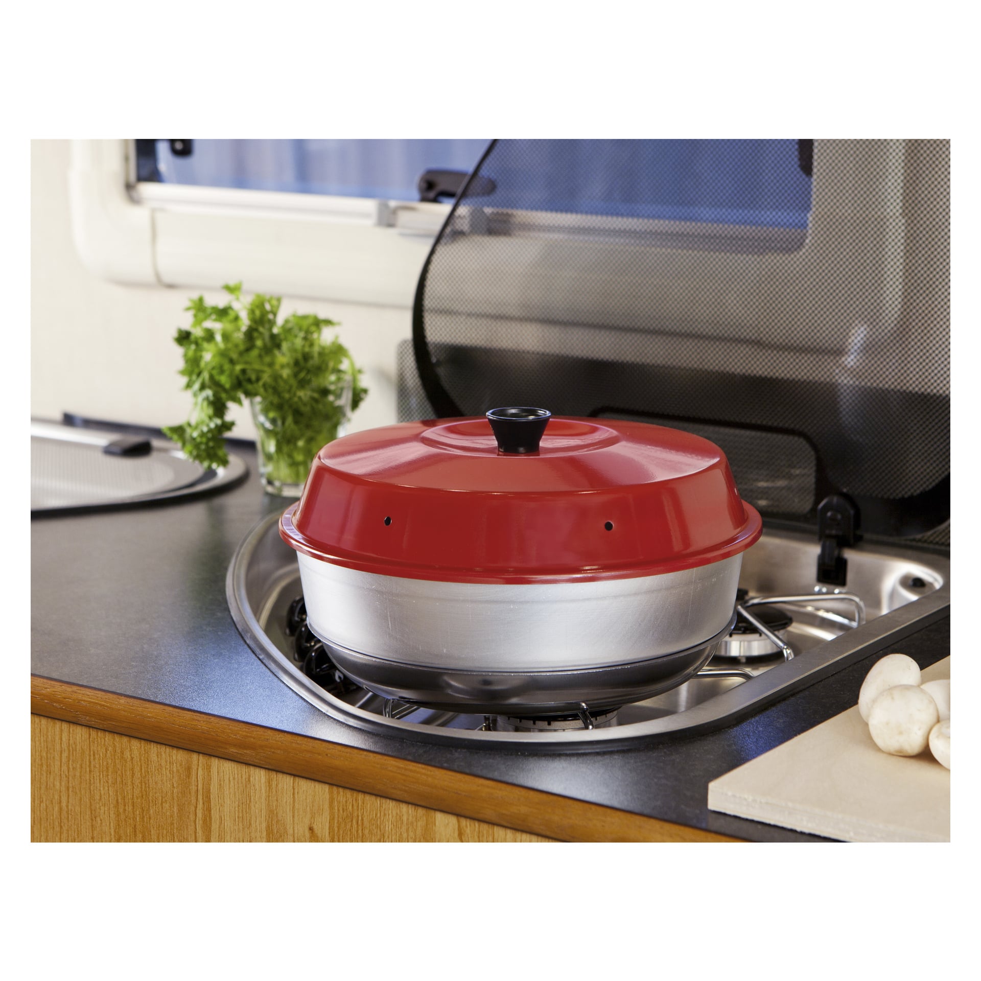 En röd och silverfärgad Omnia Cooker-ugn står på en gasplatta i köket, med färsk persilja synlig bakom och svampar arrangerade på en skärbräda i närheten.