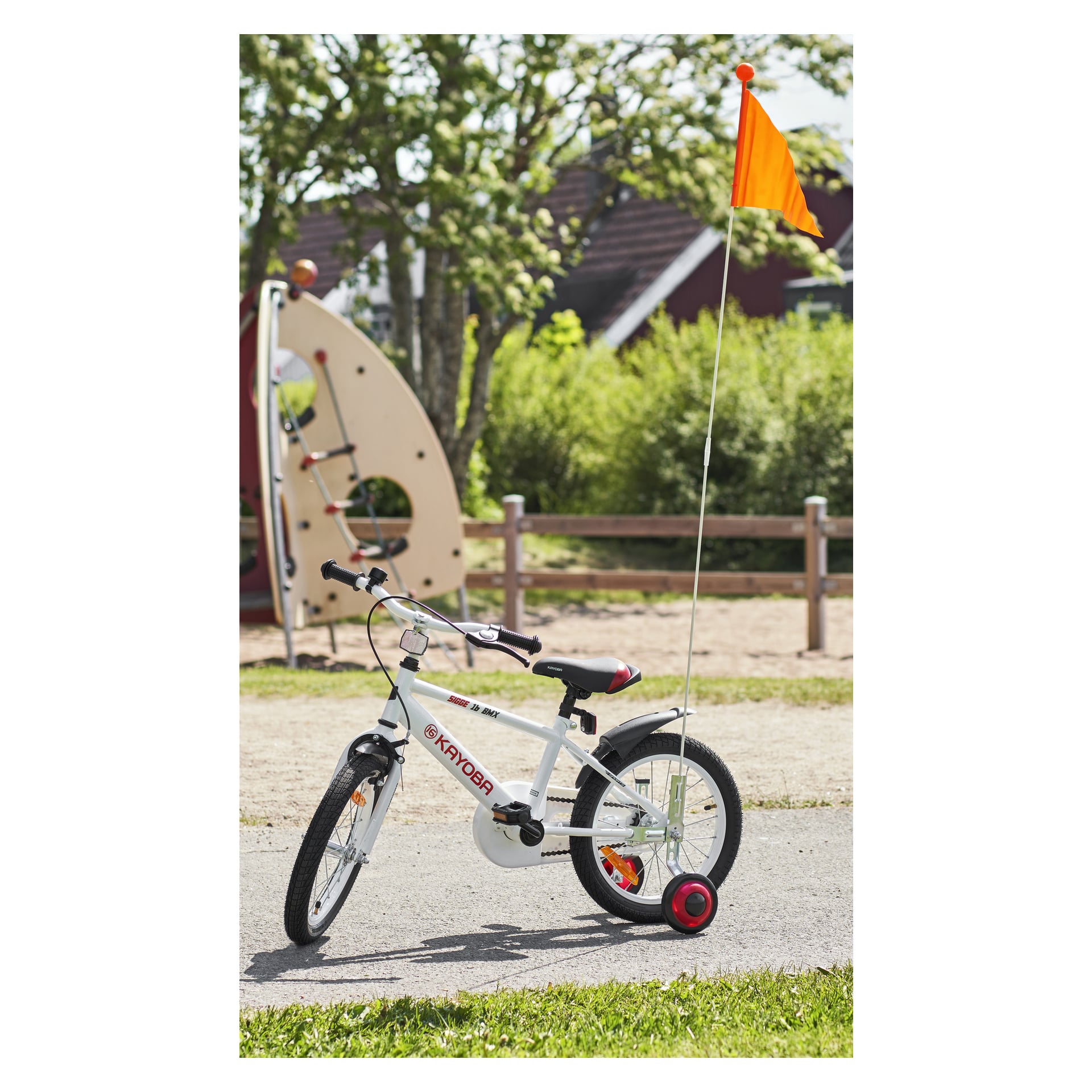 En liten vit barncykel med stödhjul och en Kayoba Bike-flagga står på en asfalterad gångväg, med färgglada lekredskap och lövgröna träd i bakgrunden.