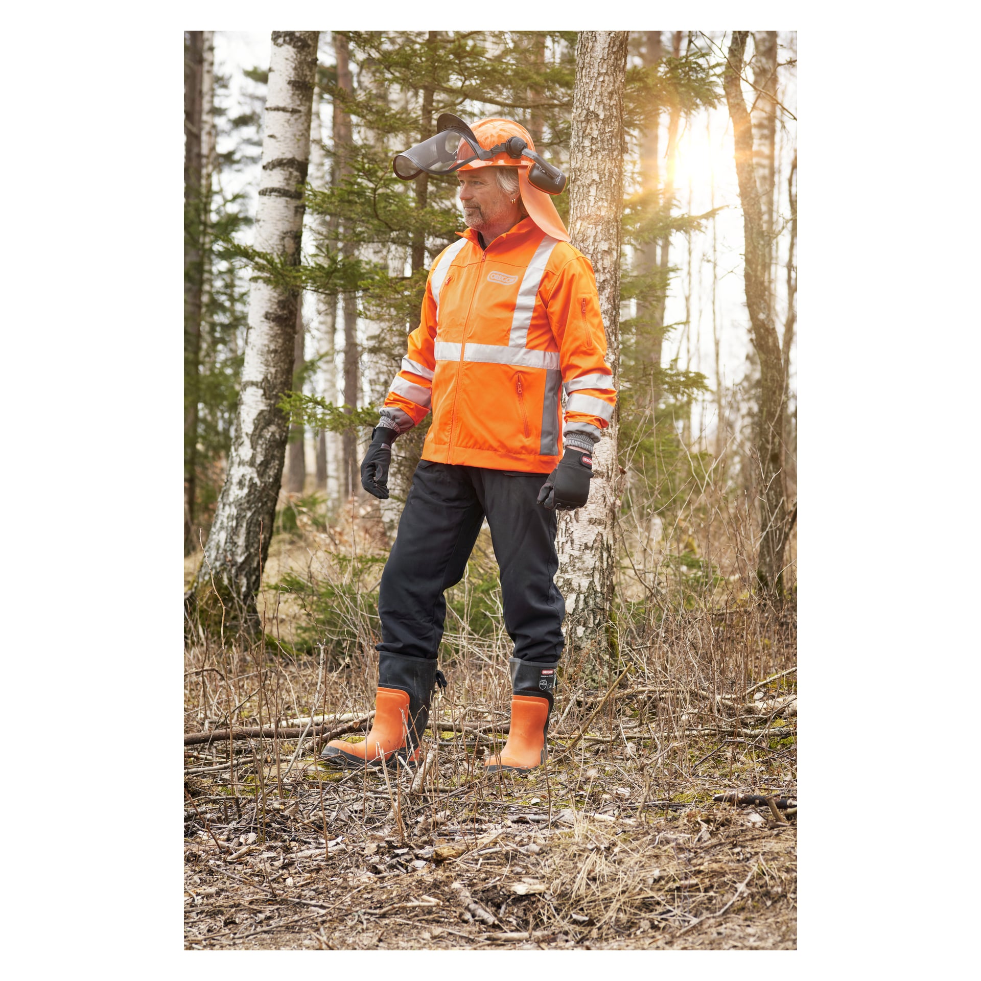 En person som bär skyddsbyxor från Oregon Saw Yukon, tillsammans med en orange skyddsjacka, matchande orange stövlar, handskar och en hjälm med ansiktsskydd, står bland solbelysta träd i en skog.