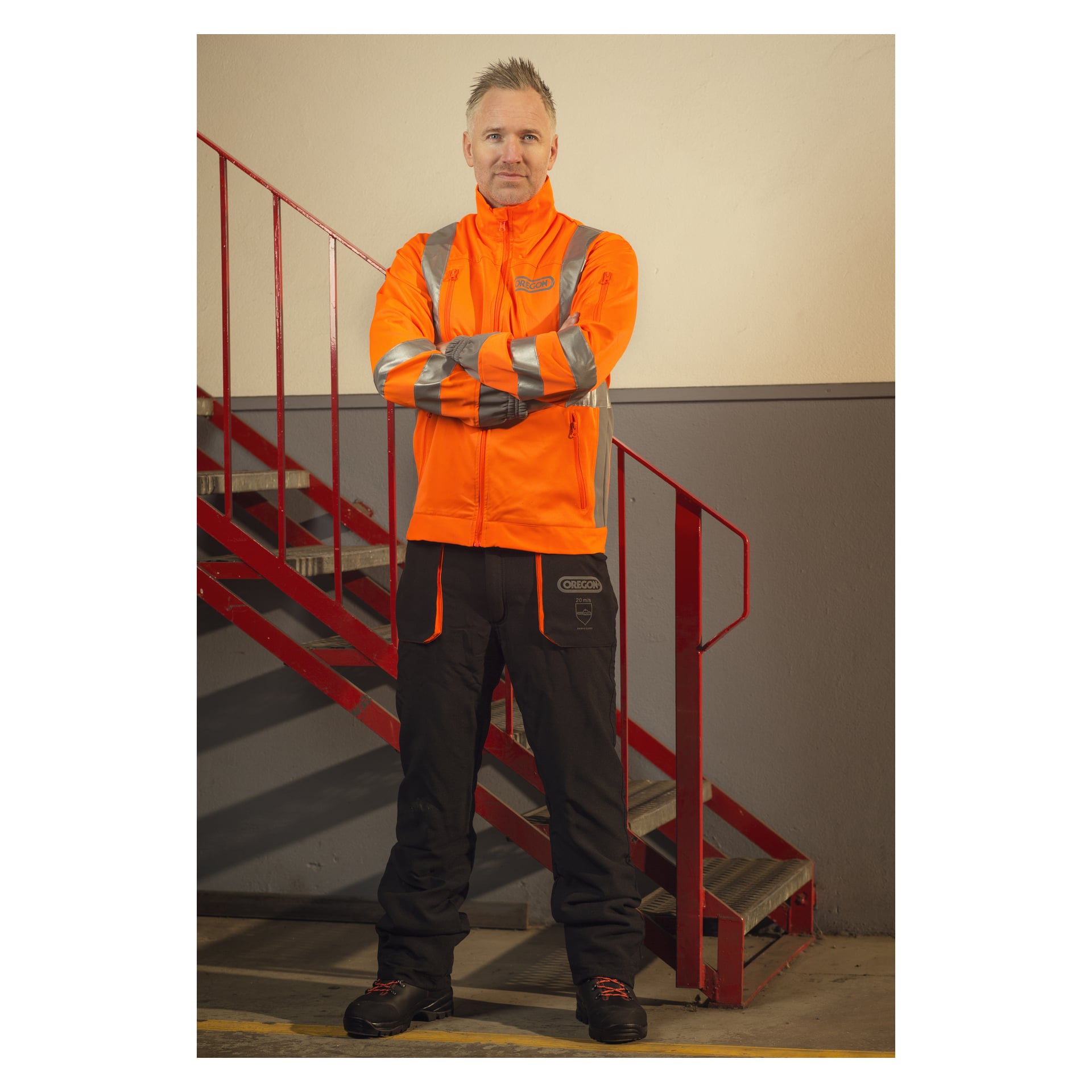 En person står inomhus med armarna i kors, klädd i en klarorange högsynlig jacka med reflekterande ränder och bär skyddsbyxor från Yukon Oregon Saw. Bakom dem finns en röd metalltrappa och en gråvit vägg.