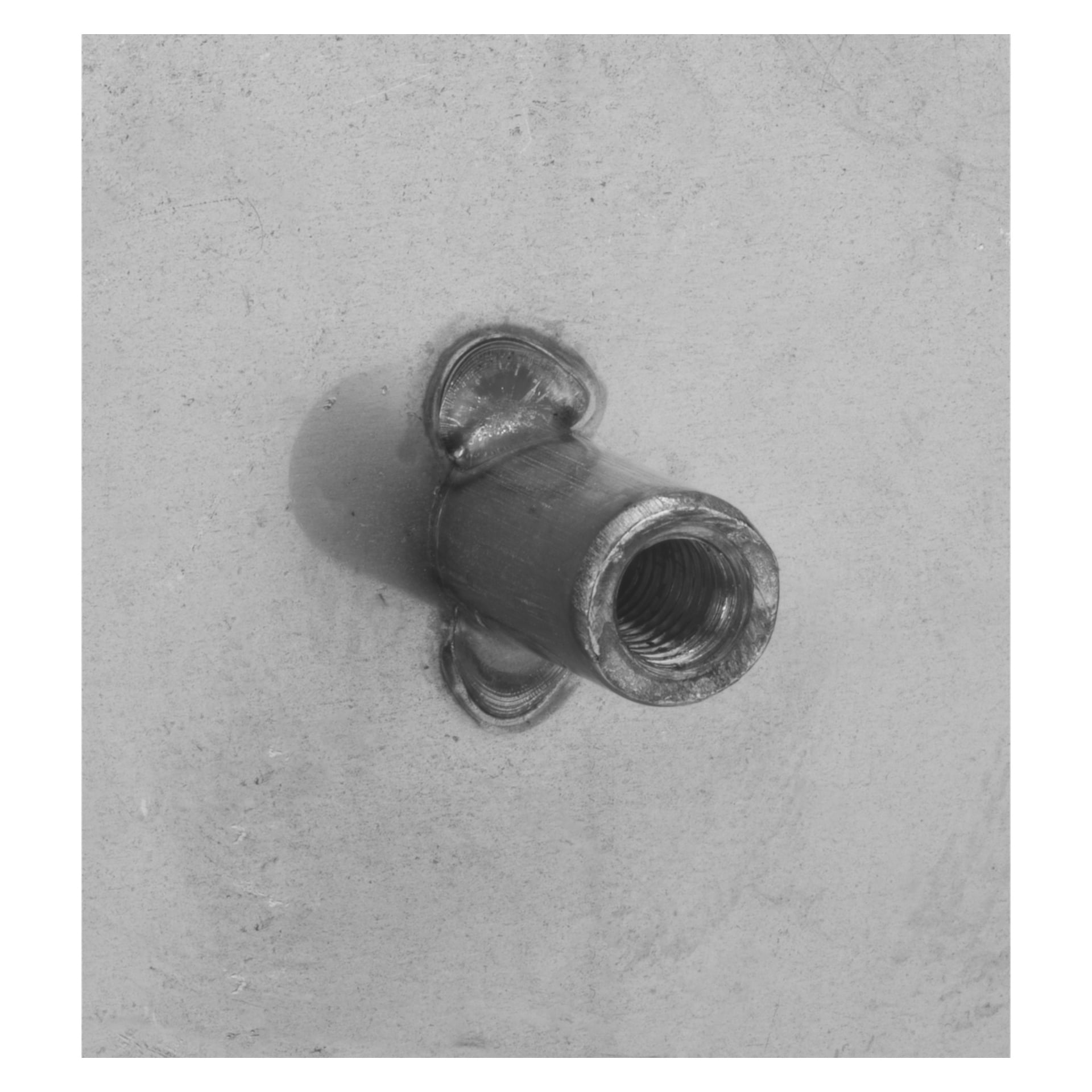 Ett Hard Head husnummernummer är fäst på en blek, plan yta. På baksidan visar metallinsatsen två svetspunkter och har en cylindrisk, öppen form riktad mot betraktaren. Fotot är i svartvitt.