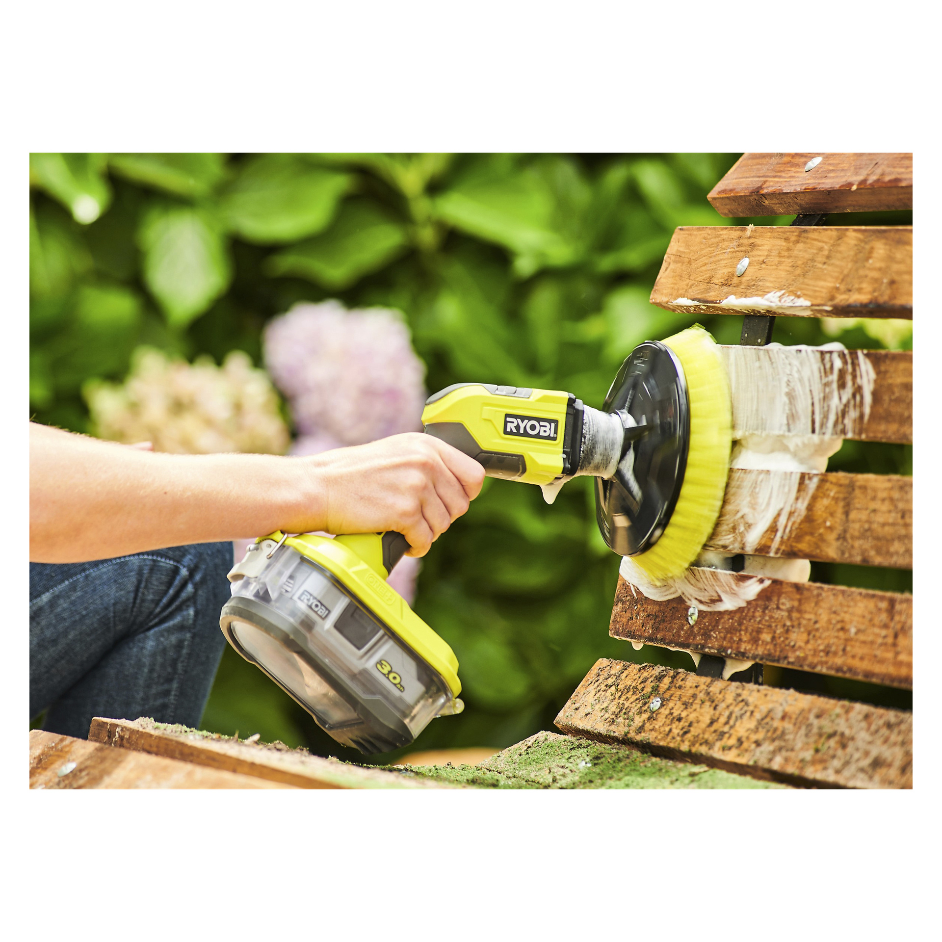Någon rengör en träbänk utomhus med en Ryobi One+ roterande tvättborste R18CPS-0 utrustad med gult borsthuvud, vilket ger tvålskum på ytan. Bakgrunden har suddig grönska och blommor.