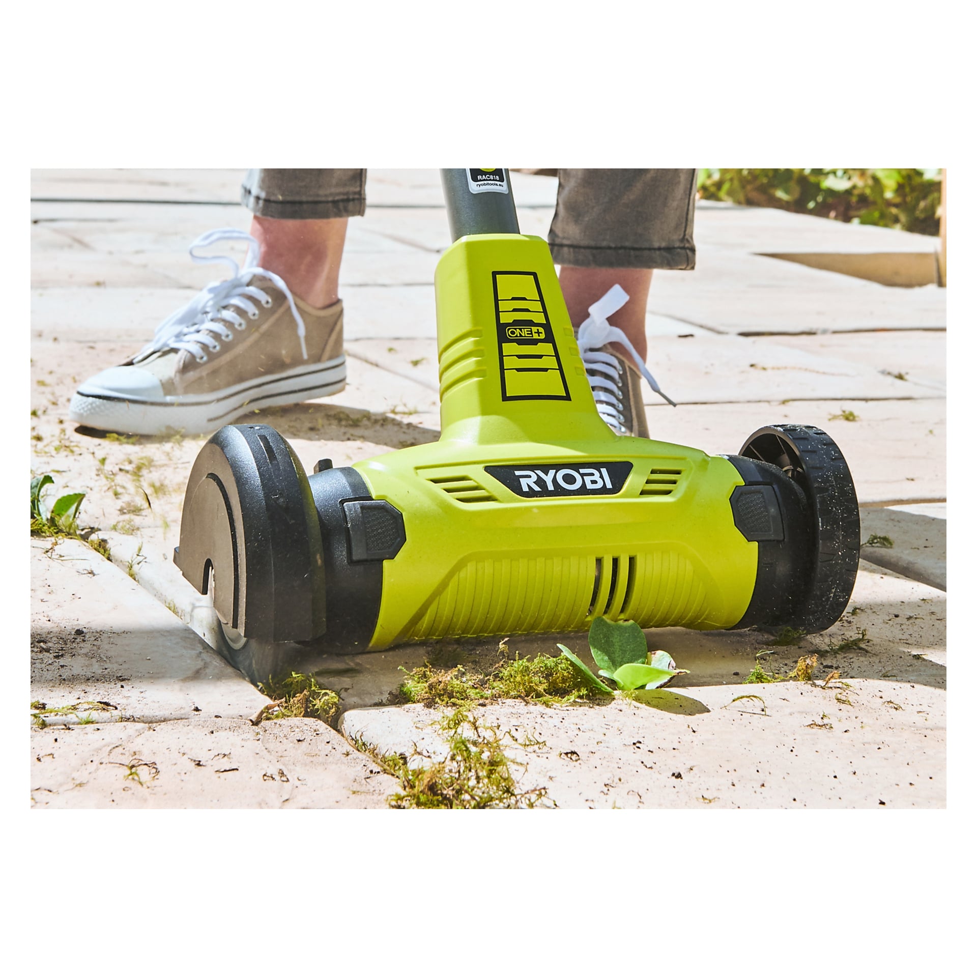 Någon tar bort ogräs och mossa mellan uteplatsstenarna med en Ryobi One+ nylonborste för att lägga skrapan RAC818, med bara underbenen och skorna synliga.