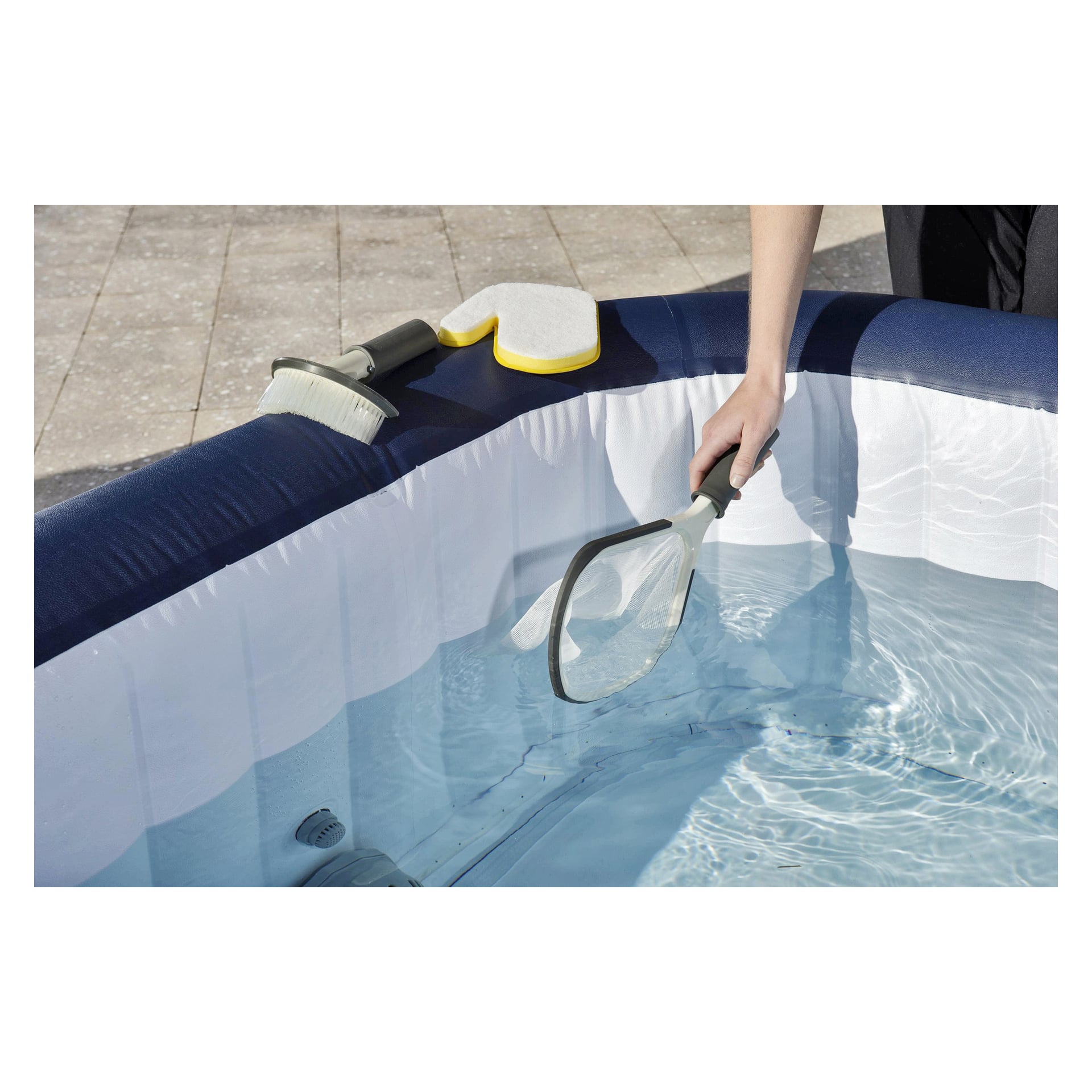 Någon rengör en uppblåsbar pool fylld med klart vatten med hjälp av det tredelade Bestway Hot Tub Cleaning Set. Borsten och svampen från scenen placeras vid poolkanten, och en kaklad uteplats syns i bakgrunden.