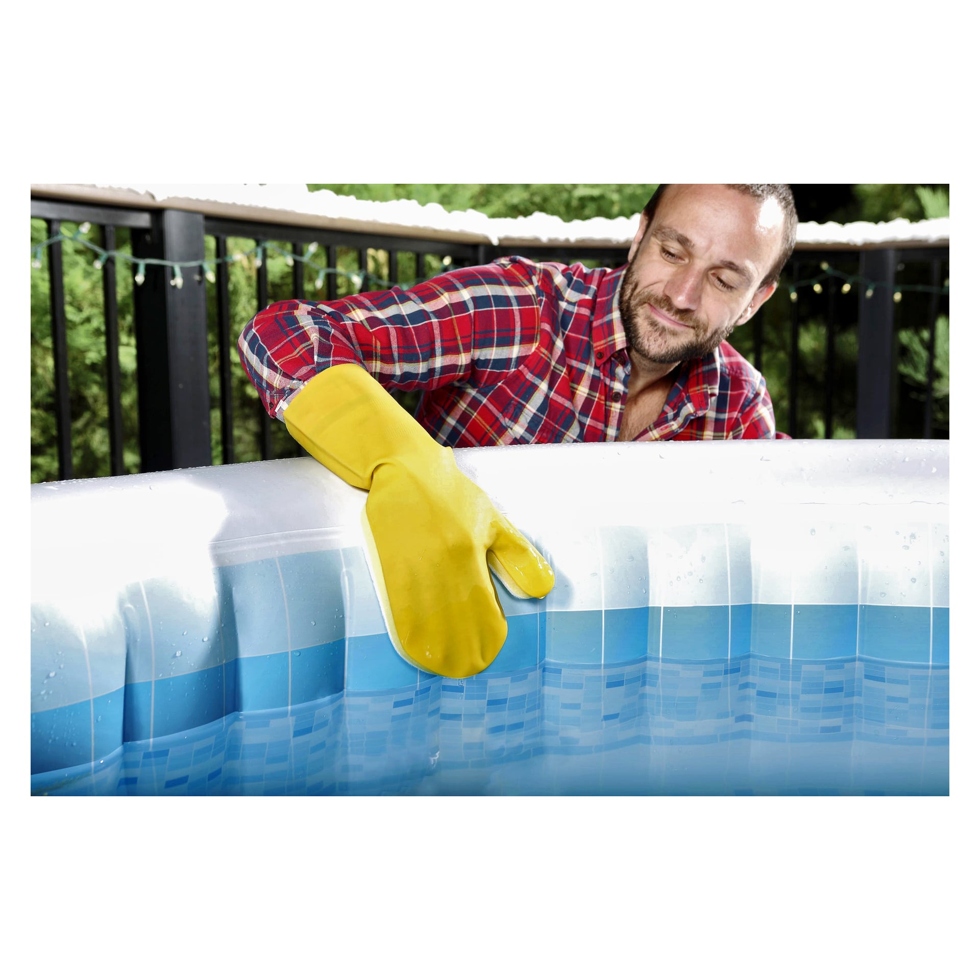 En man i en rödrutig skjorta och gula rengöringshandskar använder Bestway Hot Tub Cleaning Set (3 delar) för att skrubba kanten på en uppblåsbar pool utomhus, med träd och ett svart metallstaket som bakgrund.