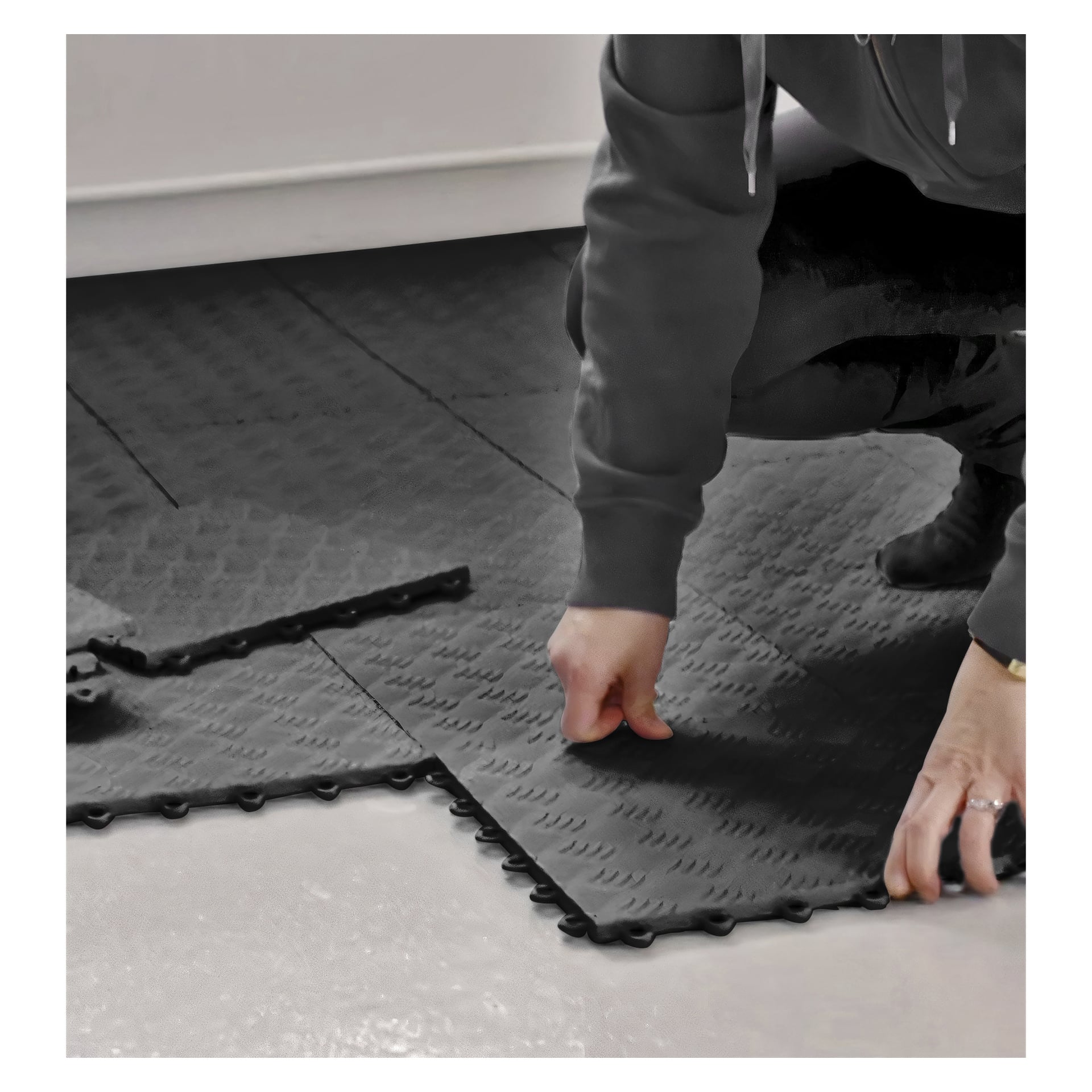 Når man kneler på gulvet, setter man sammen sammenlåste svarte skumfliser fra Hard Head Floor-fliser 9-pakningen. Bildet viser bare armen og deler av overkroppen, mens flere fliser allerede ligger på et blekt gulv.