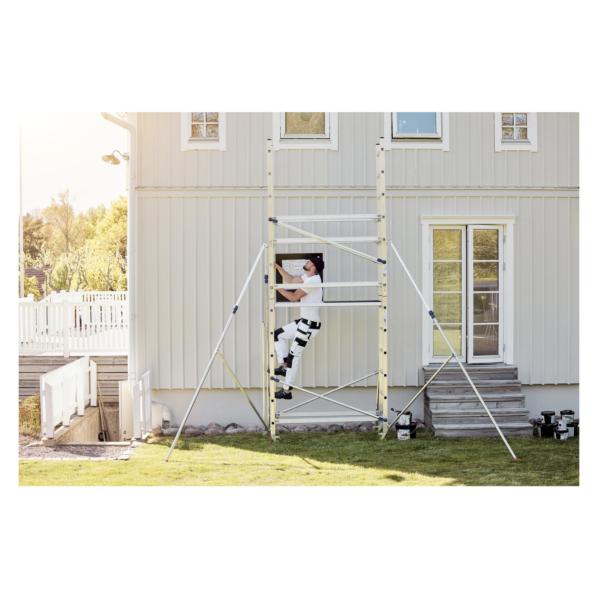 En person klädd i vitt klättrar upp på en Facas-ställning som står intill ett ljusgrått hus, med färgburkar i närheten och solljus som lyser upp området.