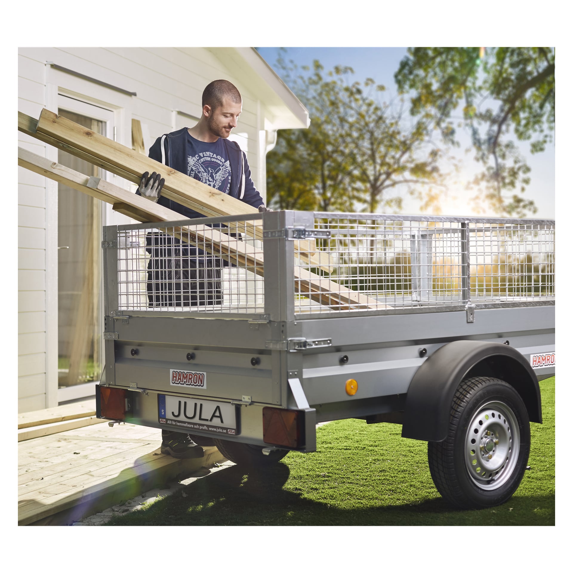 En person står bredvid ett vitt hus och placerar träplankor på en metalltrailer med nätsidor. Husvagnen står på gräsbevuxen mark nära ett träd under en ljus, solig himmel.