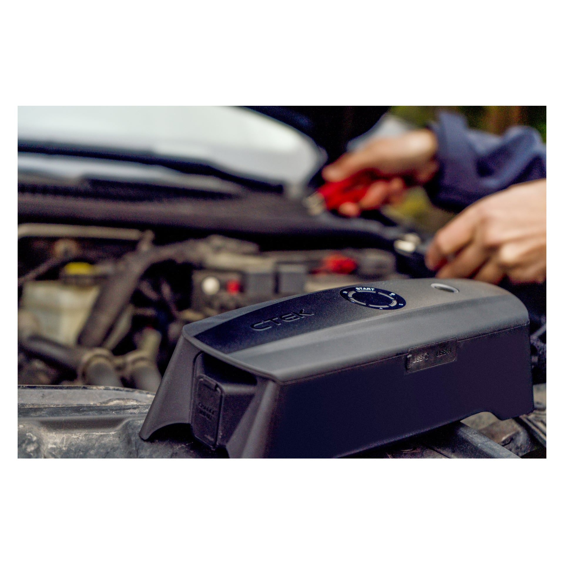 En CTEK batterilader CS Free sitter oppå en bilmotor, med en person i bakgrunnen som jobber med verktøy med rødt håndtak, noe som foreslår bilvedlikehold eller batterilading.