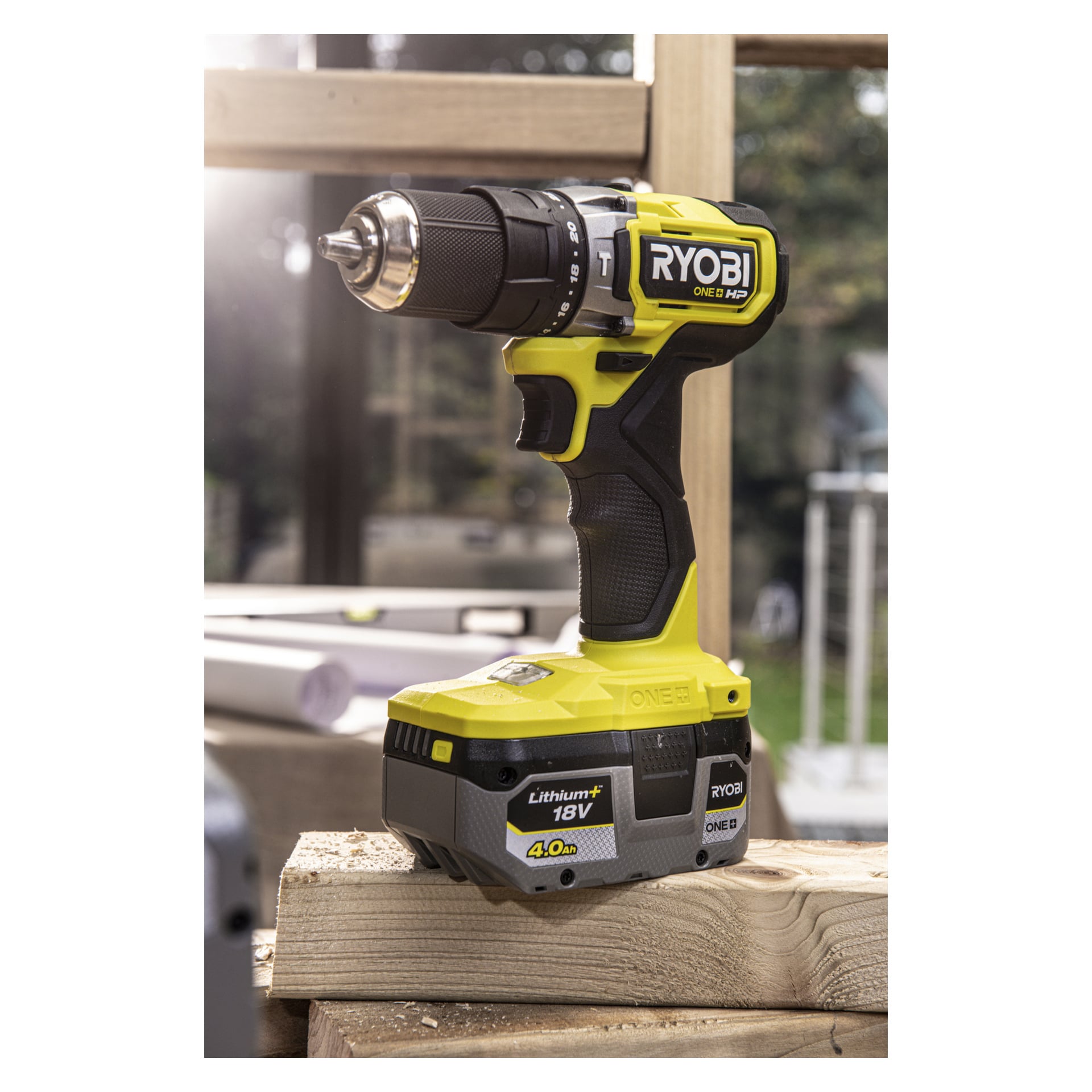 En svart och grön Ryobi One+ RB1840X 18V sladdlös borr med ett 4,0Ah-batteri står på staplade träplankor i en väl upplyst verkstad, Ryobi One+-märkningen är tydligt synlig.