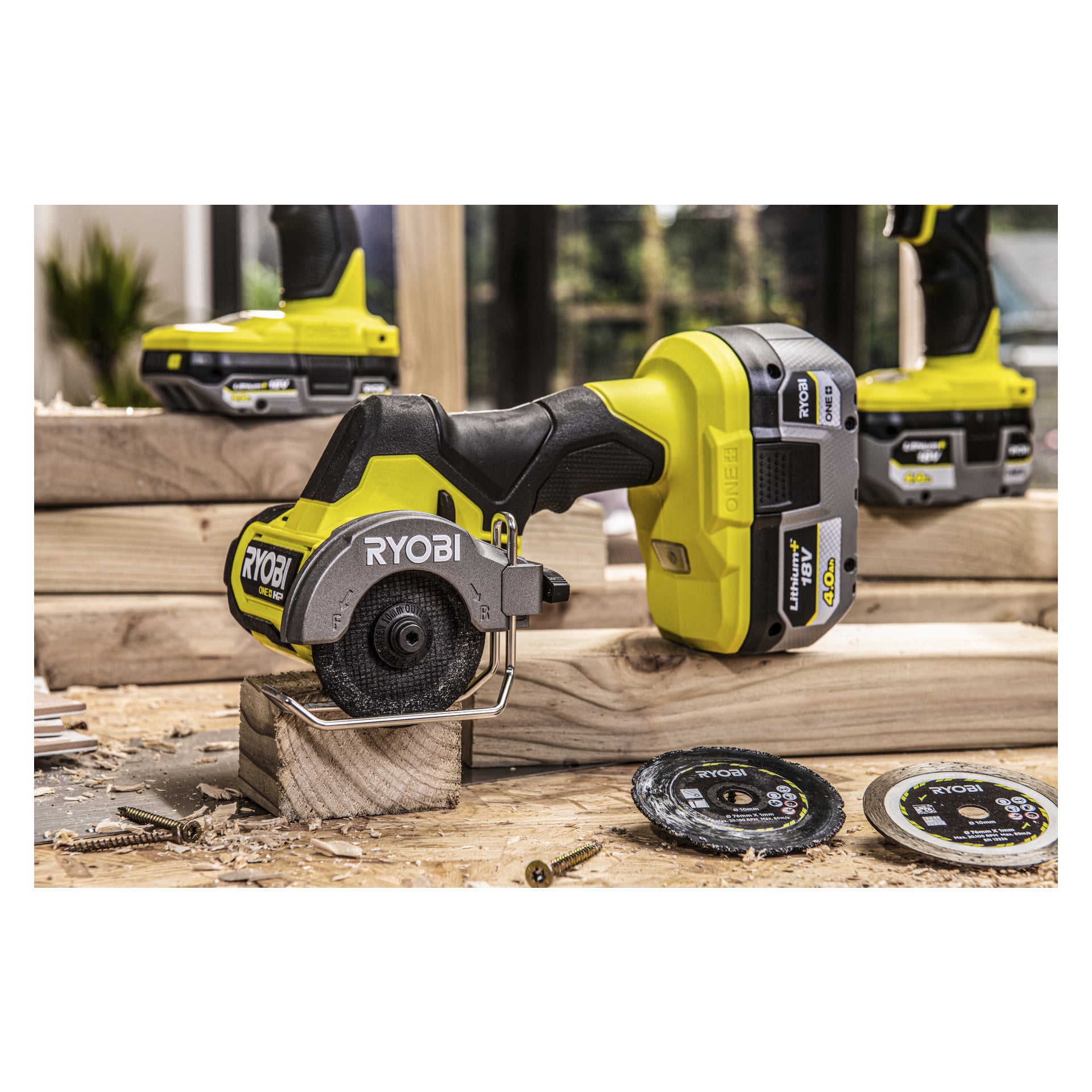 En Ryobi One+ sladdlös kraftskärare med sitt cirkulära blad visas skära genom en liten träkloss på en arbetsbänk. På samma yta finns ett RB1840X batteri, flera extra blad, skruvar och ett annat Ryobi One+-verktyg i närheten.