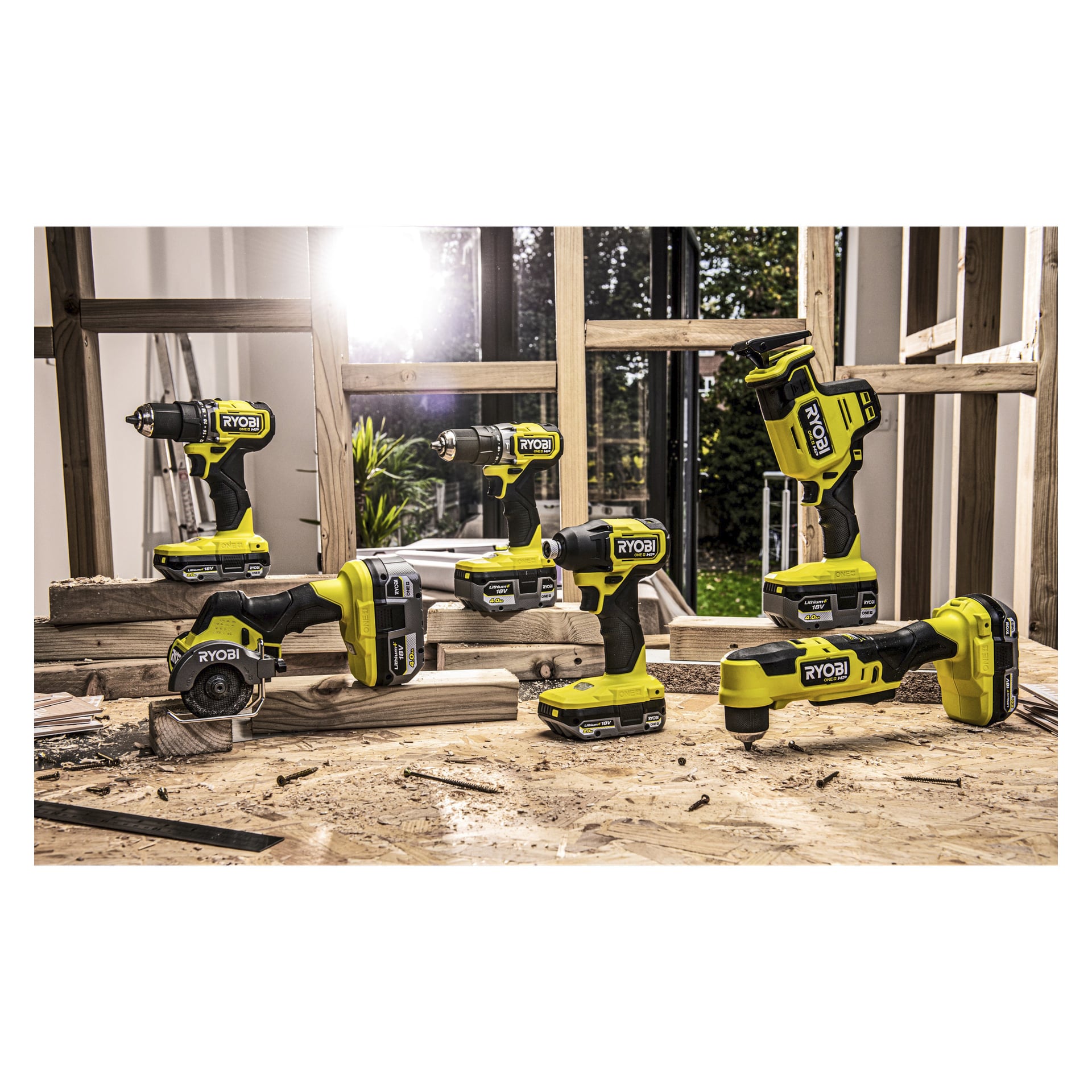 Sex Ryobi One+ Battery RB1840X-enheter står på en träarbetsbänk i ett rum under konstruktion, med stora fönster. Solljus strömmar in och framhäver batterierna och olika byggmaterial utspridda över bordet och golvet.