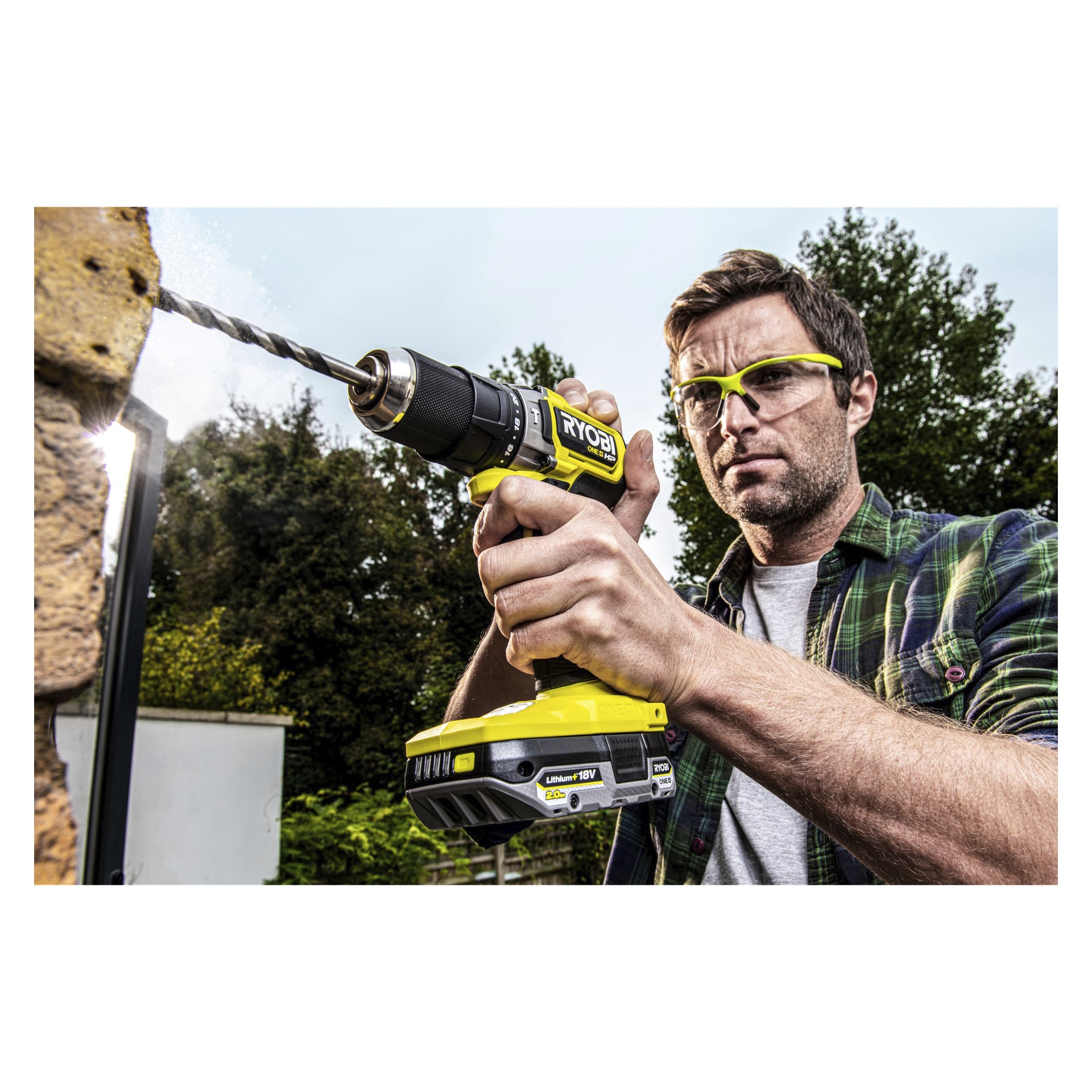 En mann med vernebriller bruker en Ryobi One+ trådløs drill drevet av et RB1820C batteri for å bore inn i en utendørs murvegg, med trær og en bygning synlig bak seg.