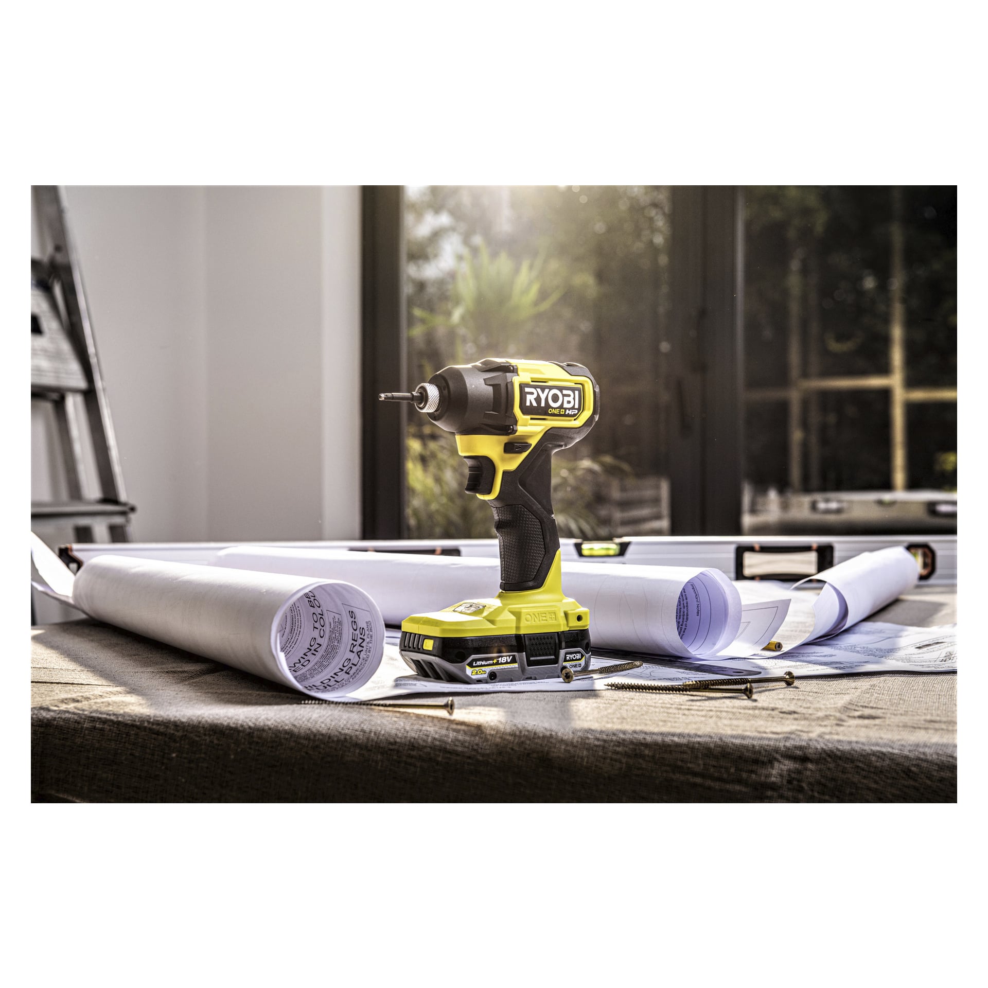 En Ryobi One+ Battery RB1820C står oppreist på et bord omgitt av sammenrullede tegninger, skruer, en blyant og et nivå, med sollys som strømmer inn gjennom store vinduer bak.