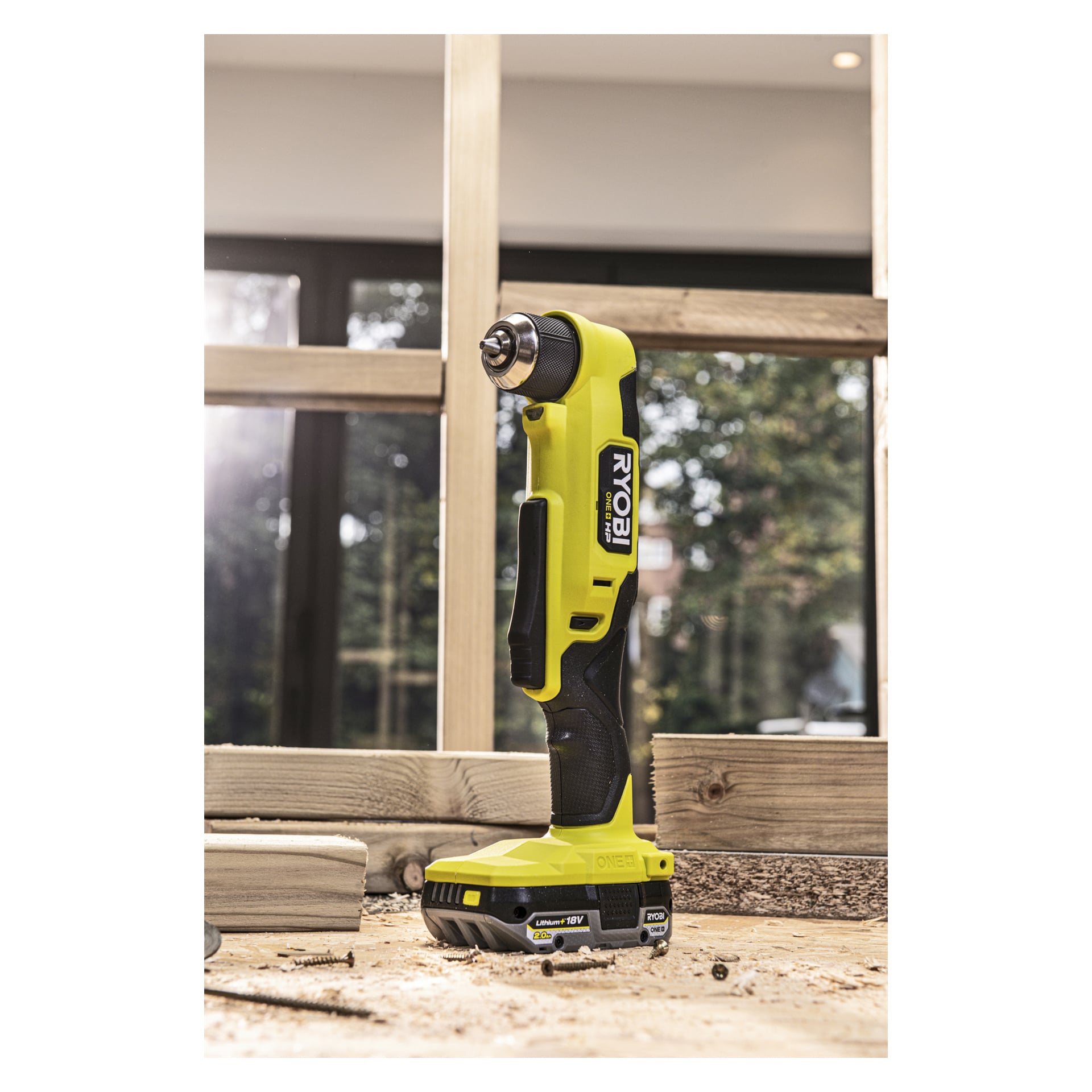 En gul og svart Ryobi One+ HP Brushless, vinklet drilldriver RAD18C-0, står oppreist på en arbeidsbenk i tre, med treflis spredt rundt og et vindu synlig i bakgrunnen.