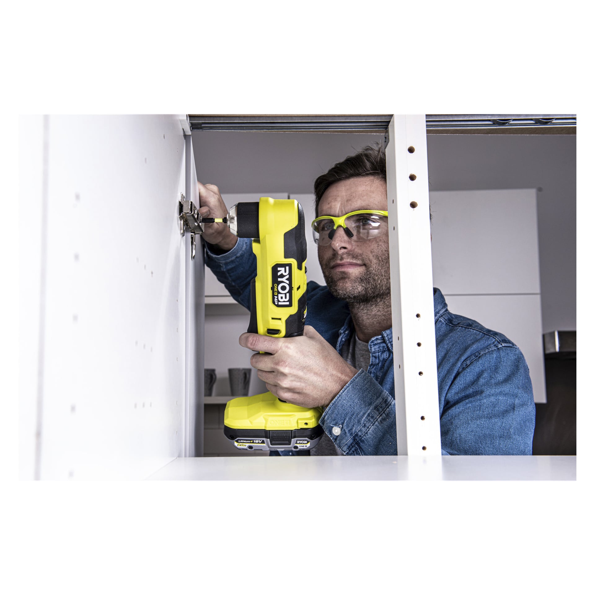 Med vernebriller bruker man Ryobi One+ HP Brushless skrå boremaskin RAD18C-0 for å feste eller justere et hengsel inne i et hvitt skap. Miljøet ligner et hjem eller verksted, med hyller og kjøkkenutstyr i bakgrunnen.