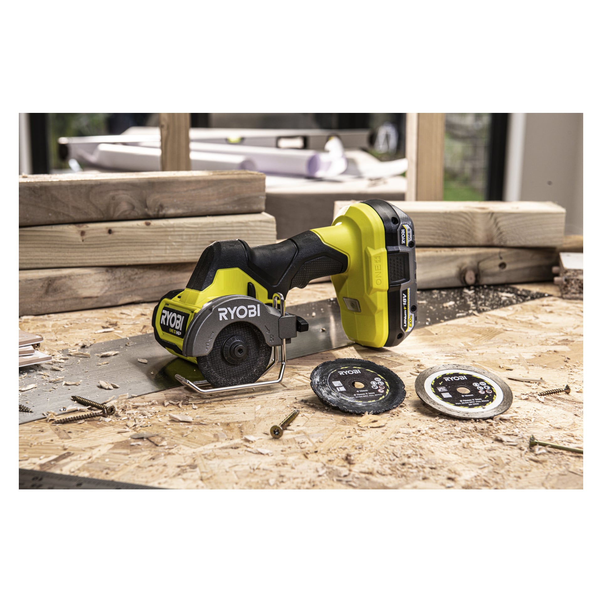 En Ryobi One+ HP Cutter RCT18C-0 batteriløs kompaktsag med svart blad hviler på en arbeidsbenk dekket med trebiter, skruer og sagflis. To ekstra Ryobi-sagblader ligger nær verktøyet.