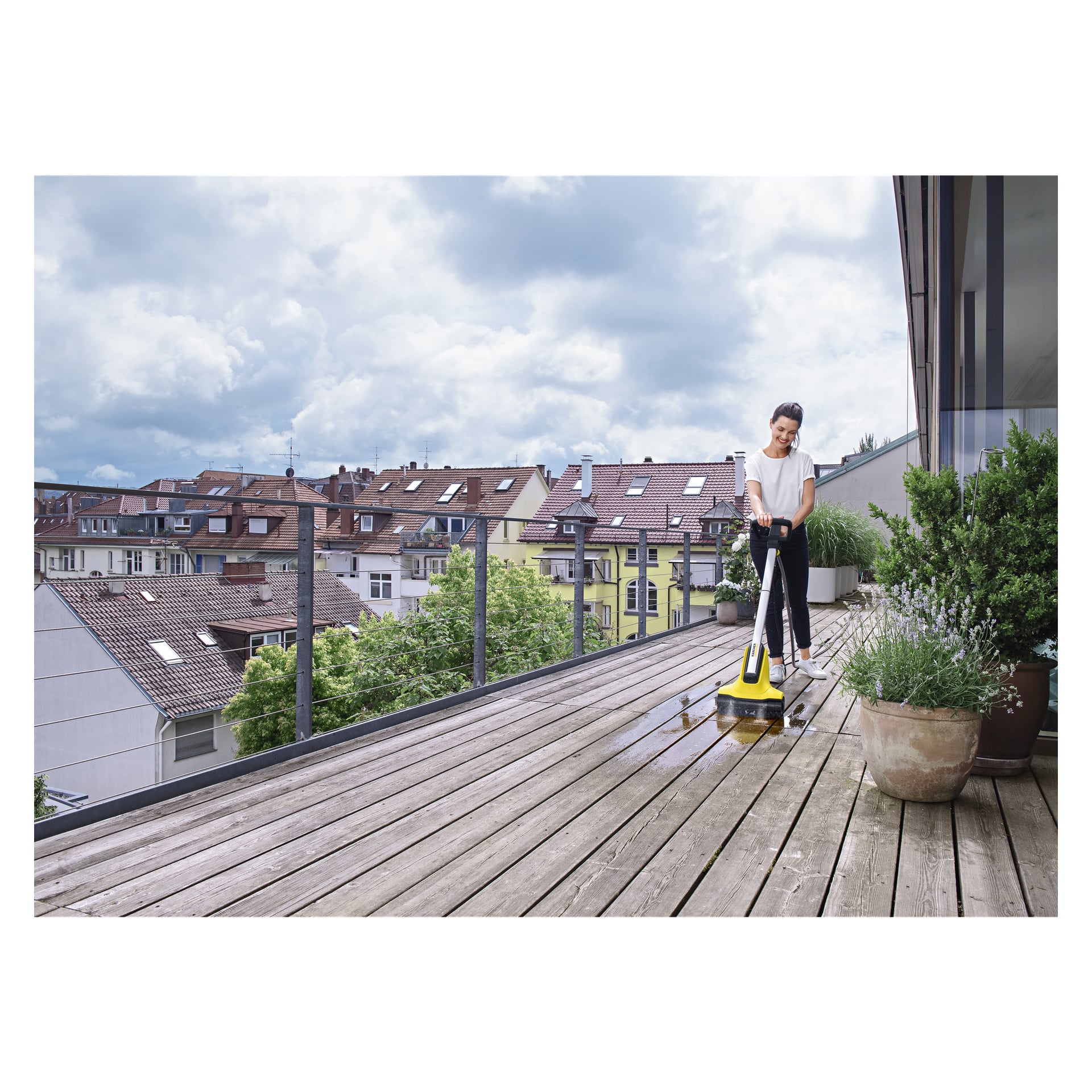 Noen rengjør en trebalkongterrasse med Kärcher Terrace wash T-Cleaner PCL4. Det er potteplanter langs den ene kanten, og bygninger med røde tak er synlige i det fjerne under en delvis skyet himmel.