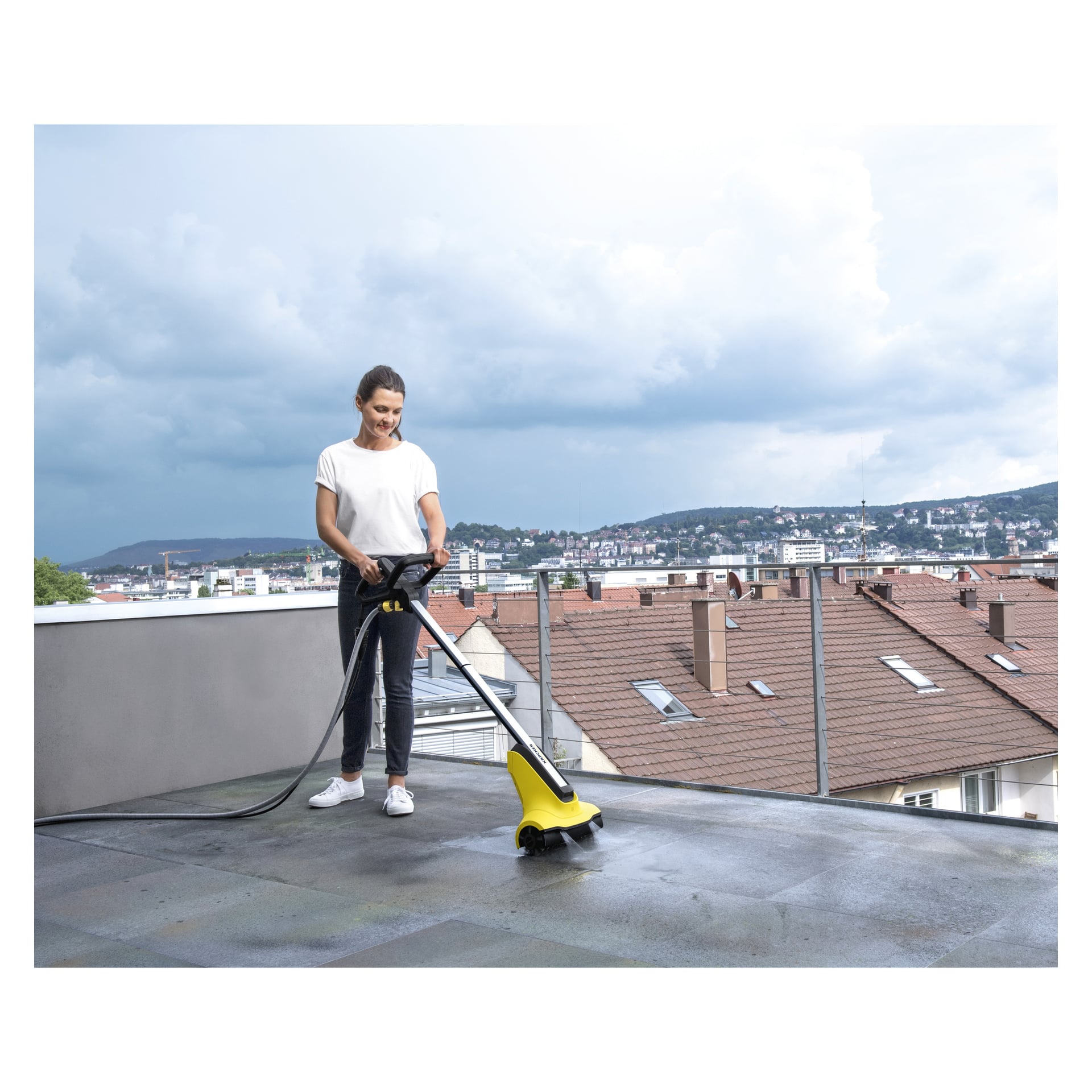 Noen kledd i hvit skjorte og mørke bukser betjener den gule og svarte Kärcher Terrace-vasken T-Cleaner PCL4 på en takterrasse, omgitt av bybygninger under en skyet himmel.