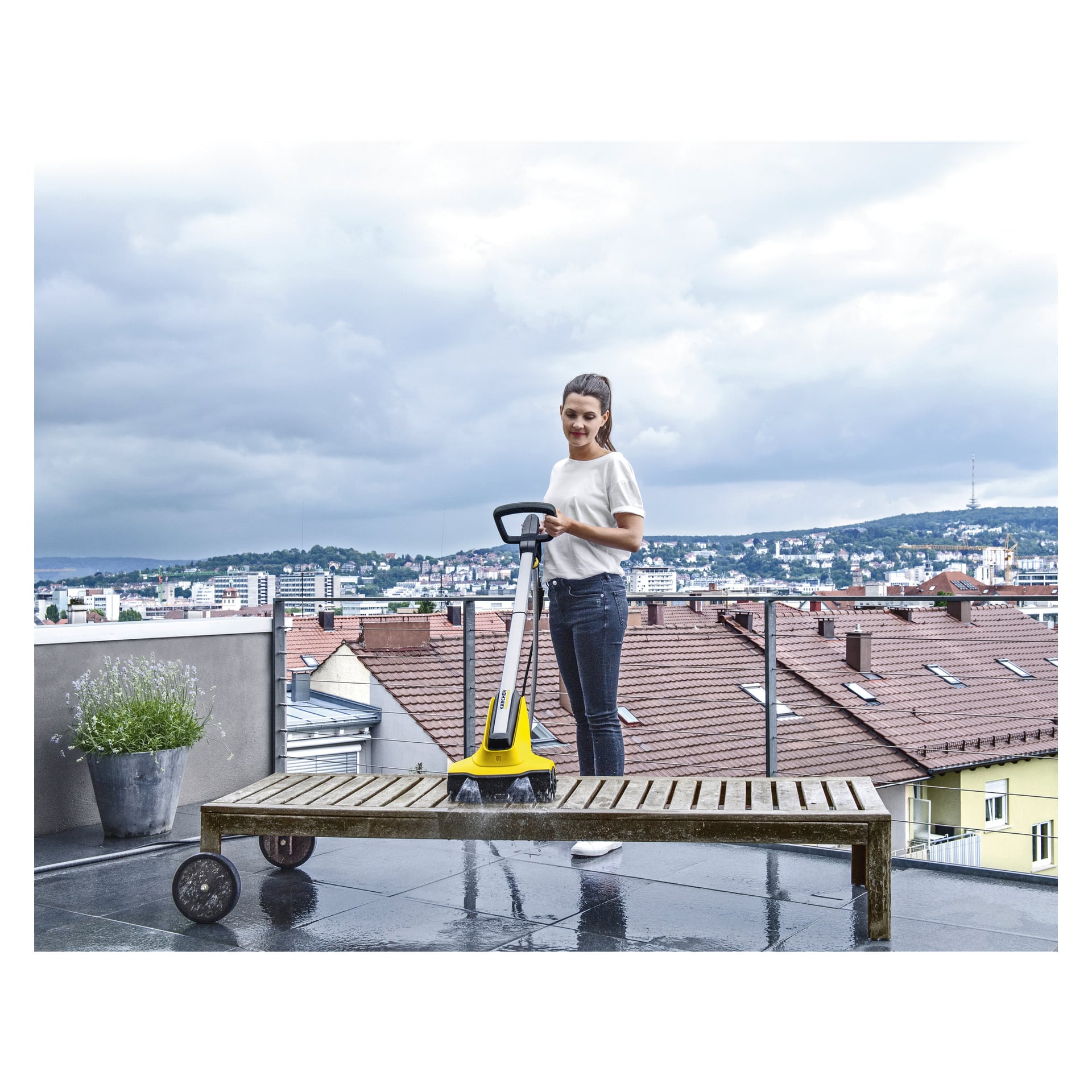 En kvinne på en balkong bruker en gul Kärcher Terrace wash T-Cleaner PCL4. I nærheten står en trebenk og en potteplante, mens bygninger og åser i det fjerne strekker seg under en overskyet himmel.