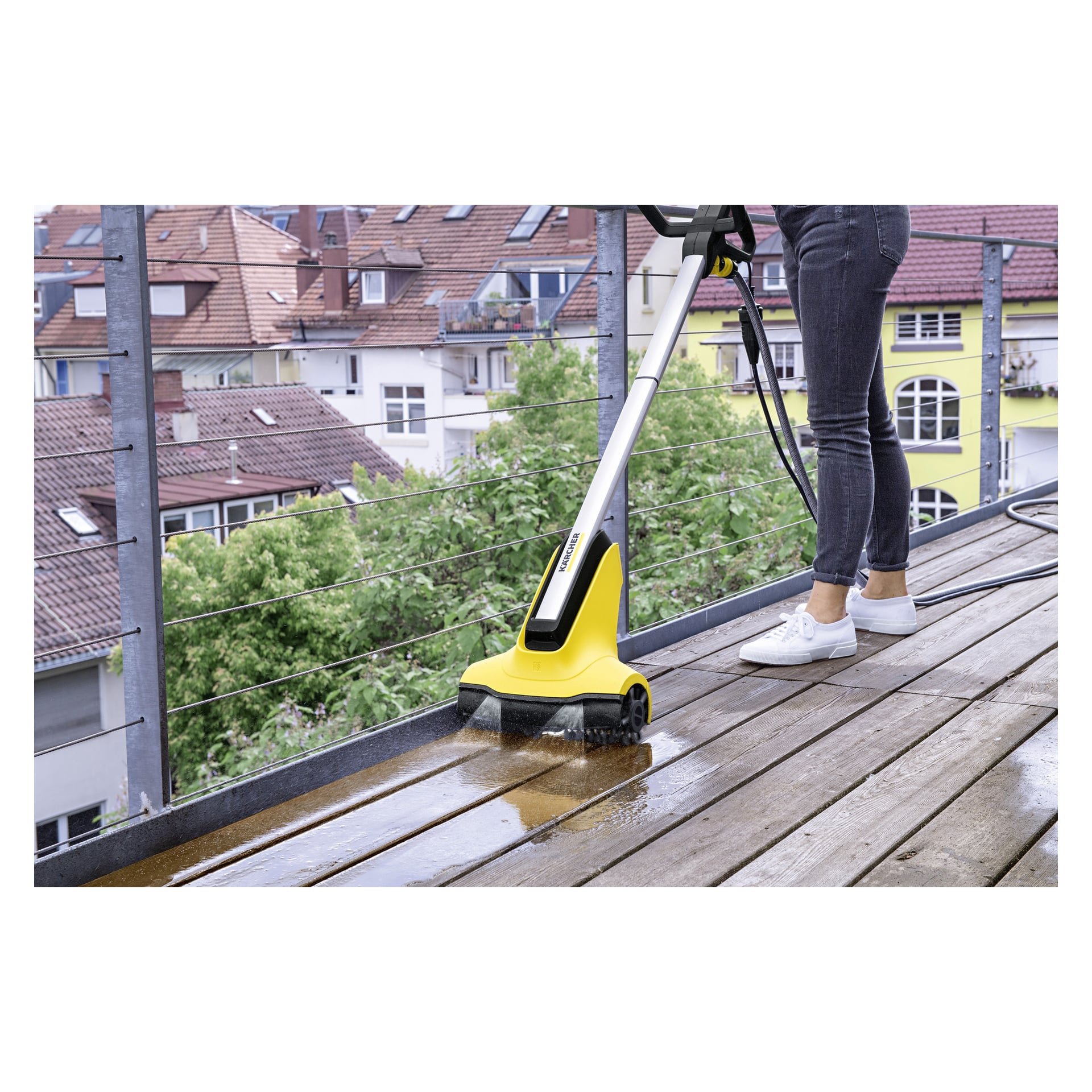 Man bruker Kärcher Terrace wash T-Cleaner PCL4 til å rengjøre en utendørs treterrasse. I bakgrunnen er det leiegårder med røde tak og grønne trær. Personen er kledd i svarte jeans og hvite joggesko.