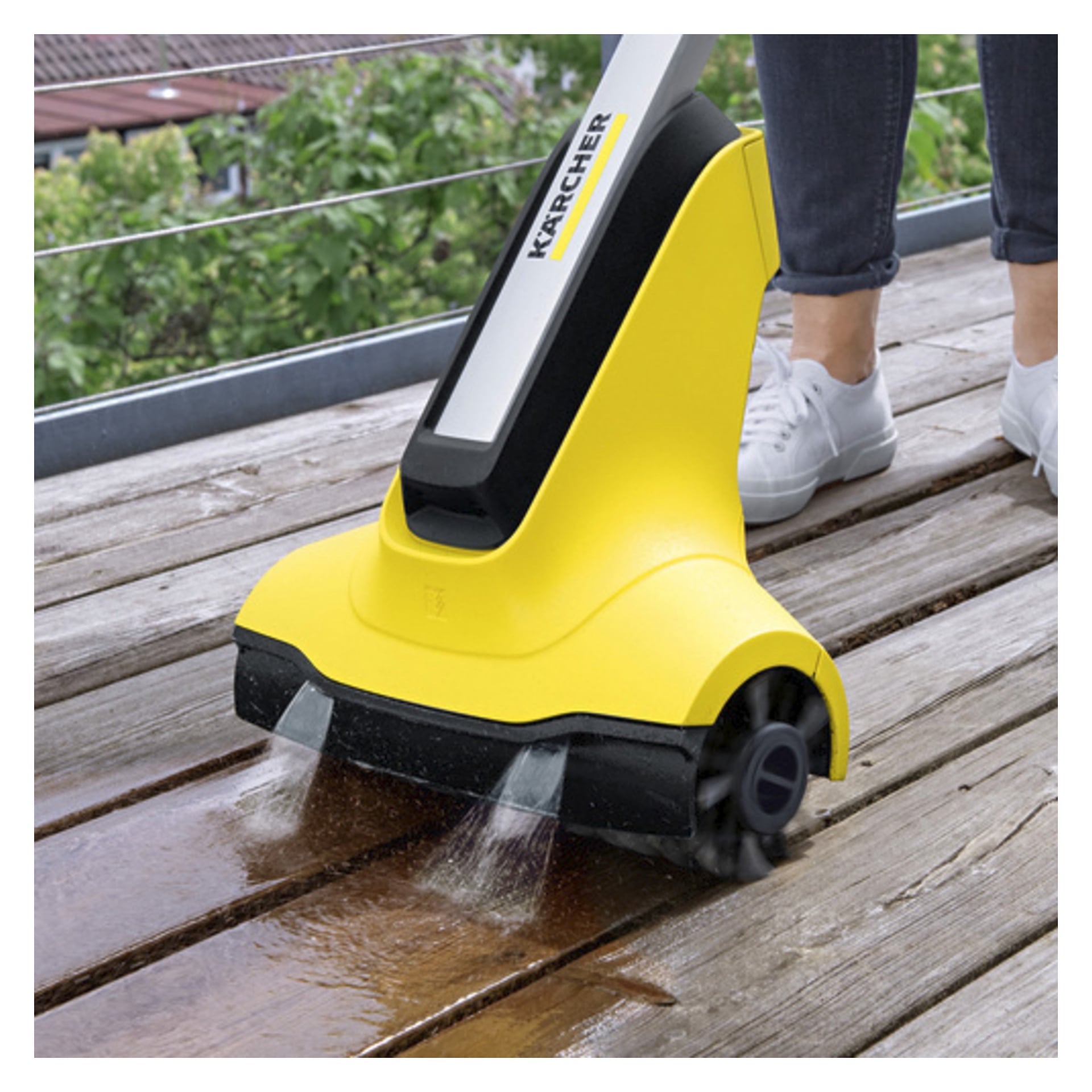 En person bruker Kärcher Terrace wash T-Cleaner PCL4 for å rengjøre et tredekk, med vannstråler som spruter fra dysene og synlig skyller treverket mens det skyves fremover.