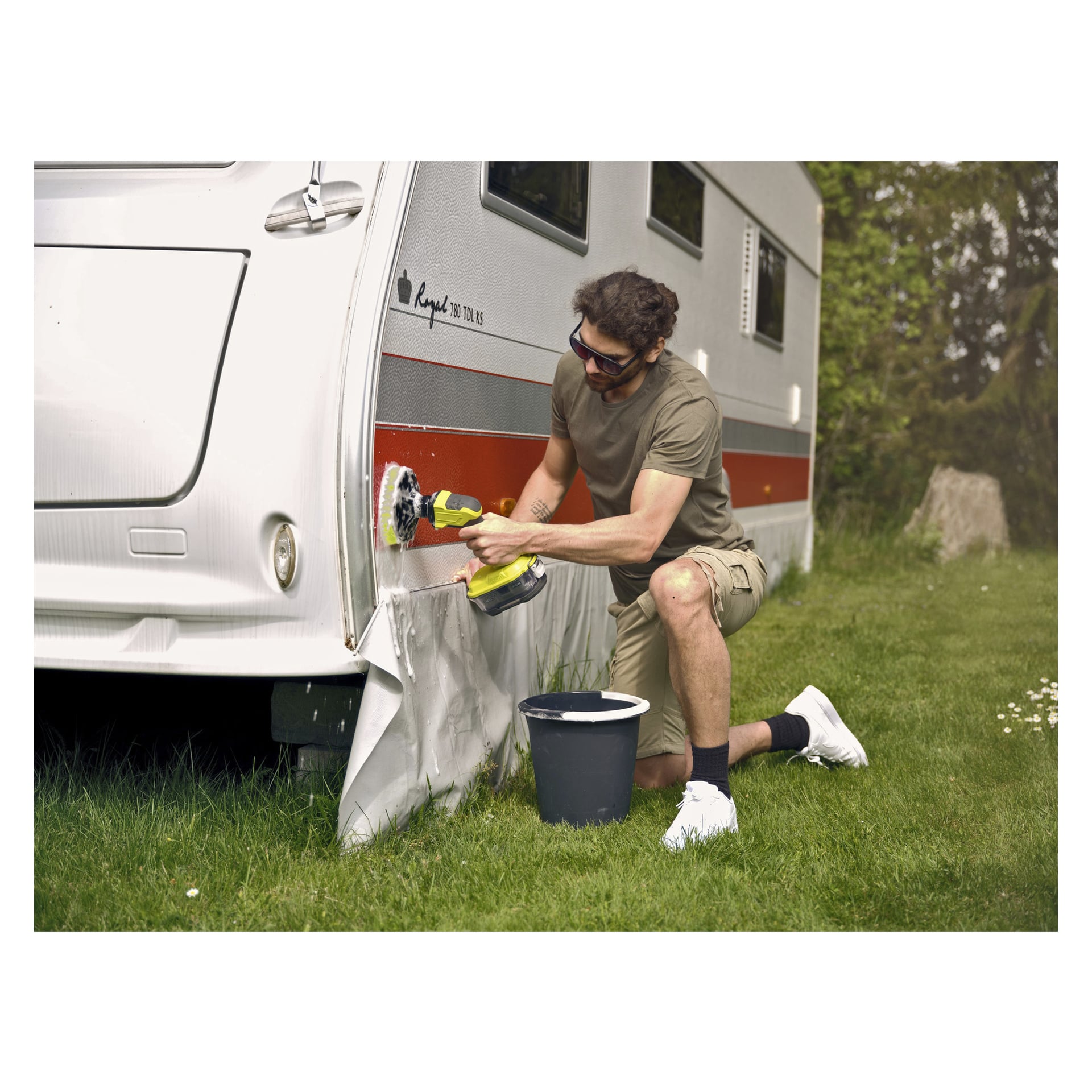 Med solglasögon, t-shirt och shorts använder man Ryobi One+ roterande tvättborste R18CPS-0 för att rengöra den nedre delen av en rödvit husbil. En svart hink står på gräset i närheten, med träd och grönska i bakgrunden.