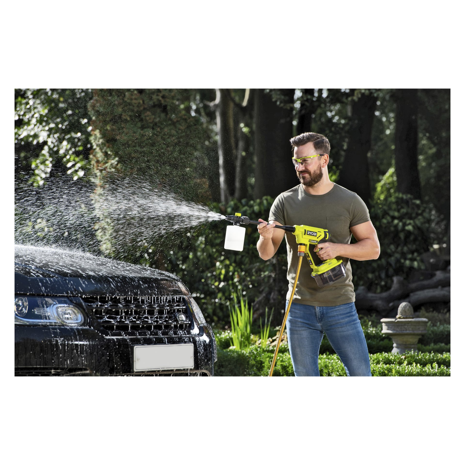 En man med skyddsglasögon använder en Ryobi One+ Chemical Container for Medium Pressure Washers RAC748 för att spraya tvåligt vatten på framsidan av en svart bil utanför, med träd och trädgårdsväxter i bakgrunden.