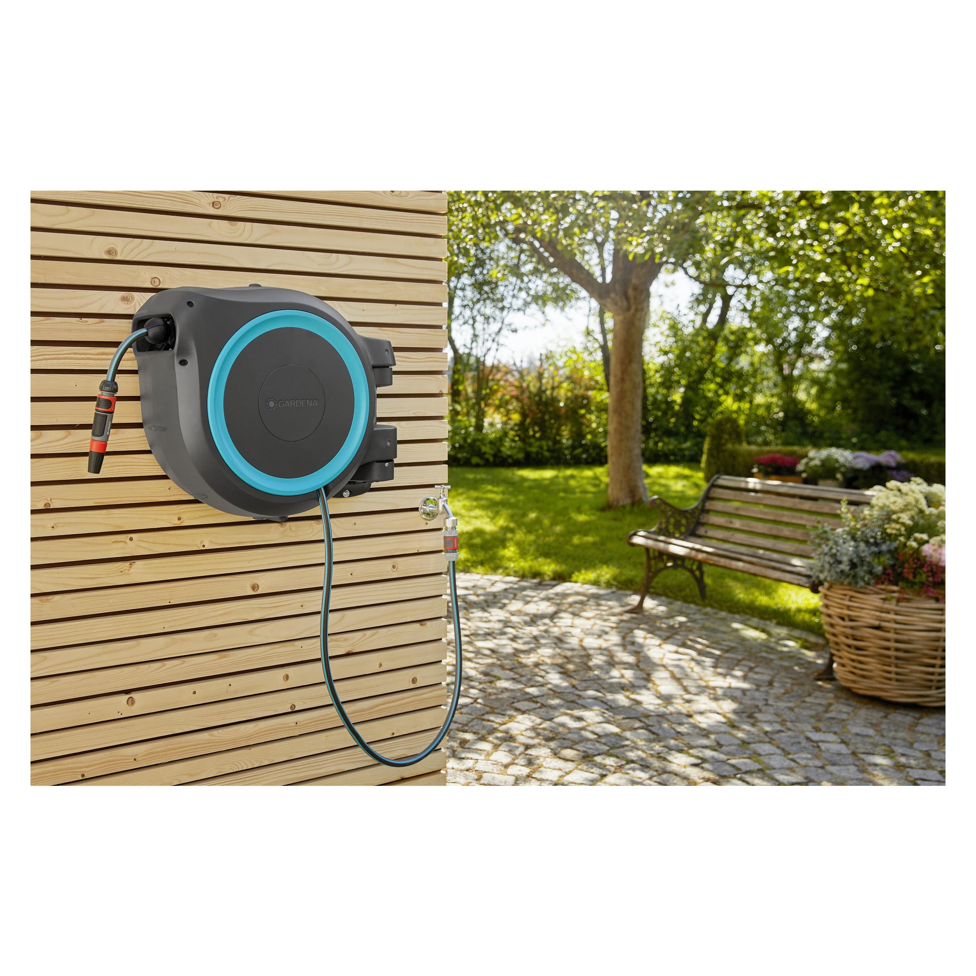 En Gardena-slangrull med vattenslang RollUp XL är fäst på en trävägg, dess slang delvis avrullad och ansluten till en vattenkran. Bakom den finns en kullerstensbelagd uteplats, en träbänk och en livfull grön trädgård.