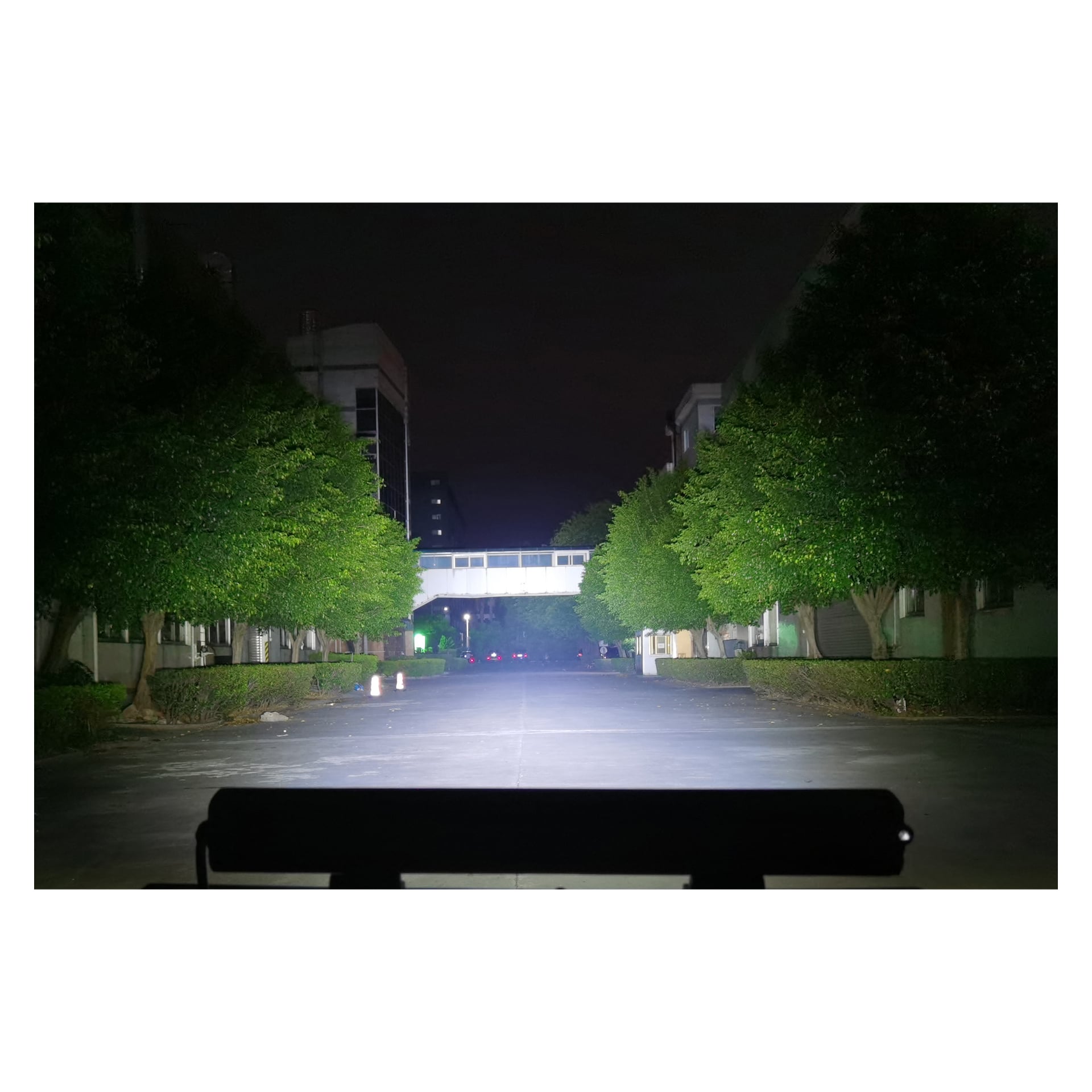 En starkt upplyst, öde gata på natten belyses av starka strålar från en Hamron LED-ljusbar. Träd flankeras på båda sidor av vägen, och en gångbro förbinder två byggnader i bakgrunden. Perspektivet är inifrån ett fordon.