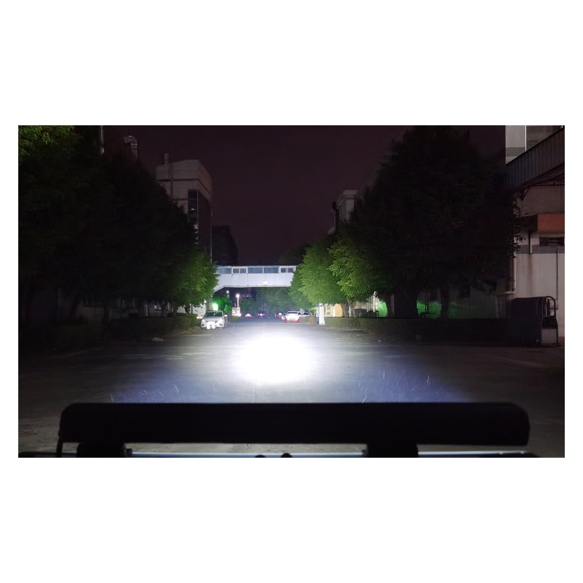 En väl upplyst gata på natten har träd på båda sidor och byggnader i bakgrunden. En Hamron LED-ljusramp i förgrunden lyser starkt upp vägen, med flera parkerade bilar under en gångbro.