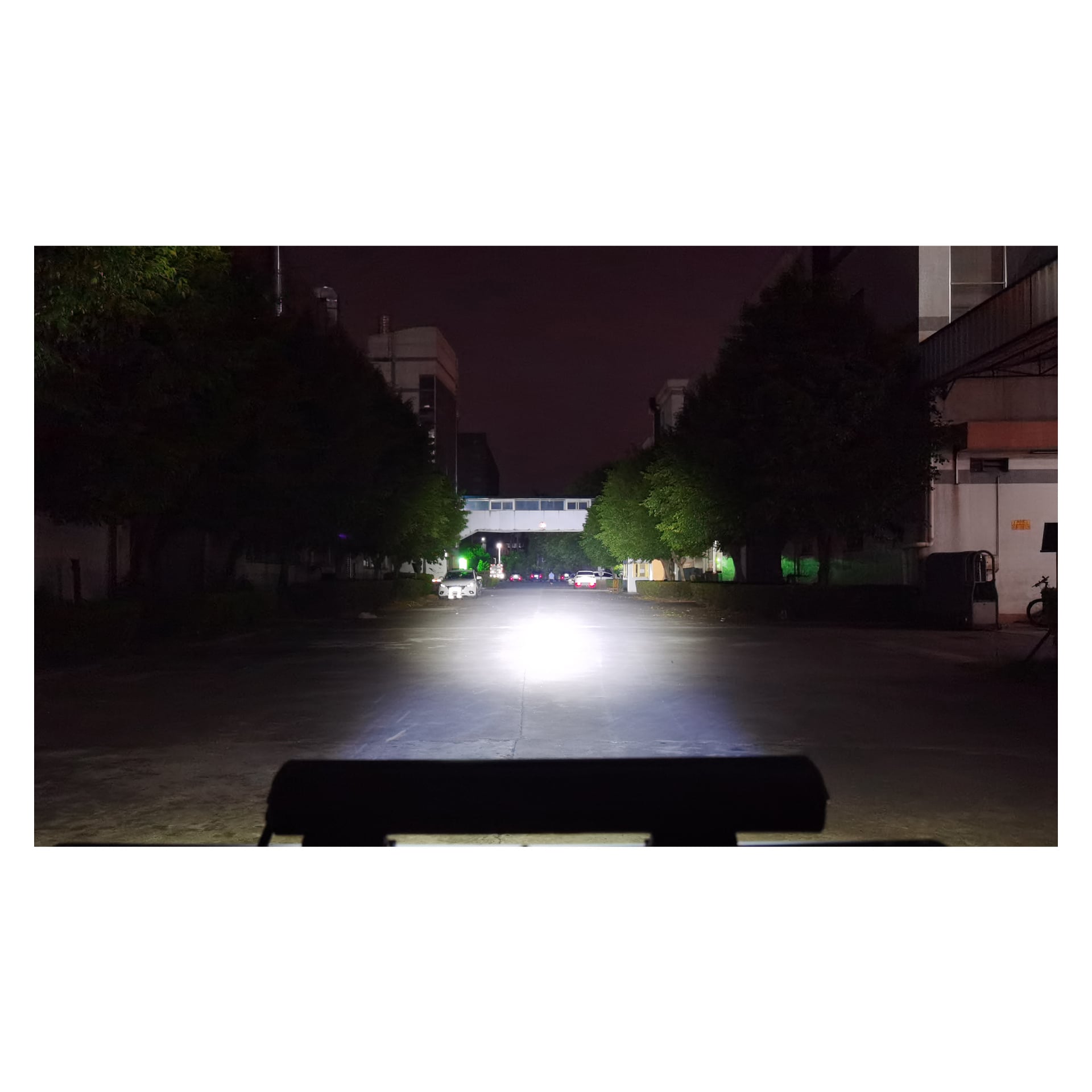 En lys Hamron Light bar LED lyser over en øde nattgate omgitt av bygninger og trær, og lyser opp noen parkerte biler og sykler under en bro i det fjerne.