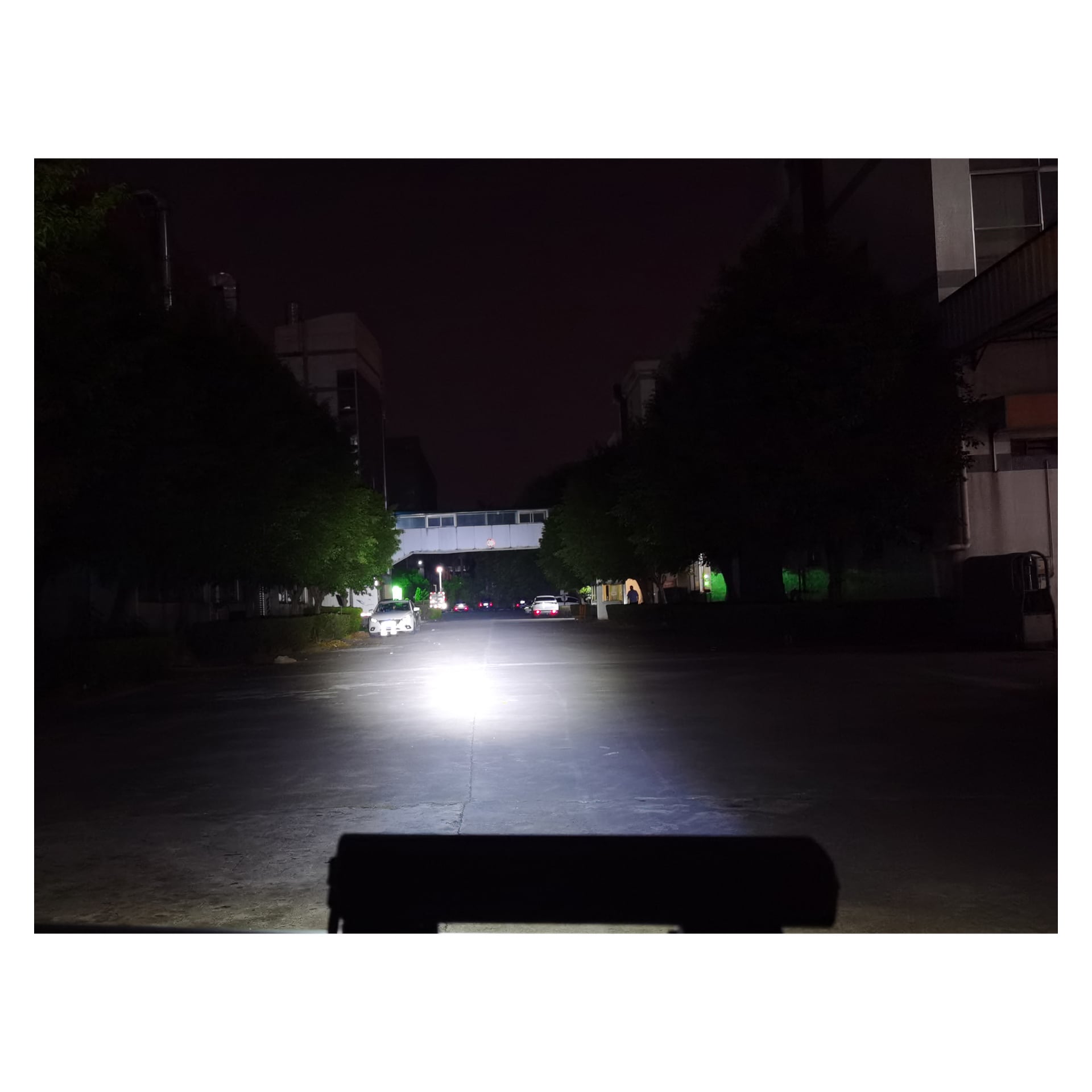 En nattlig gatescene er opplyst i forgrunnen av en LED-frontlykt med Hamron Lightbar. Biler er synlige lenger nedover veien, som passerer under en gangbro som forbinder bygninger, mens trær grenser til hver side av gaten.