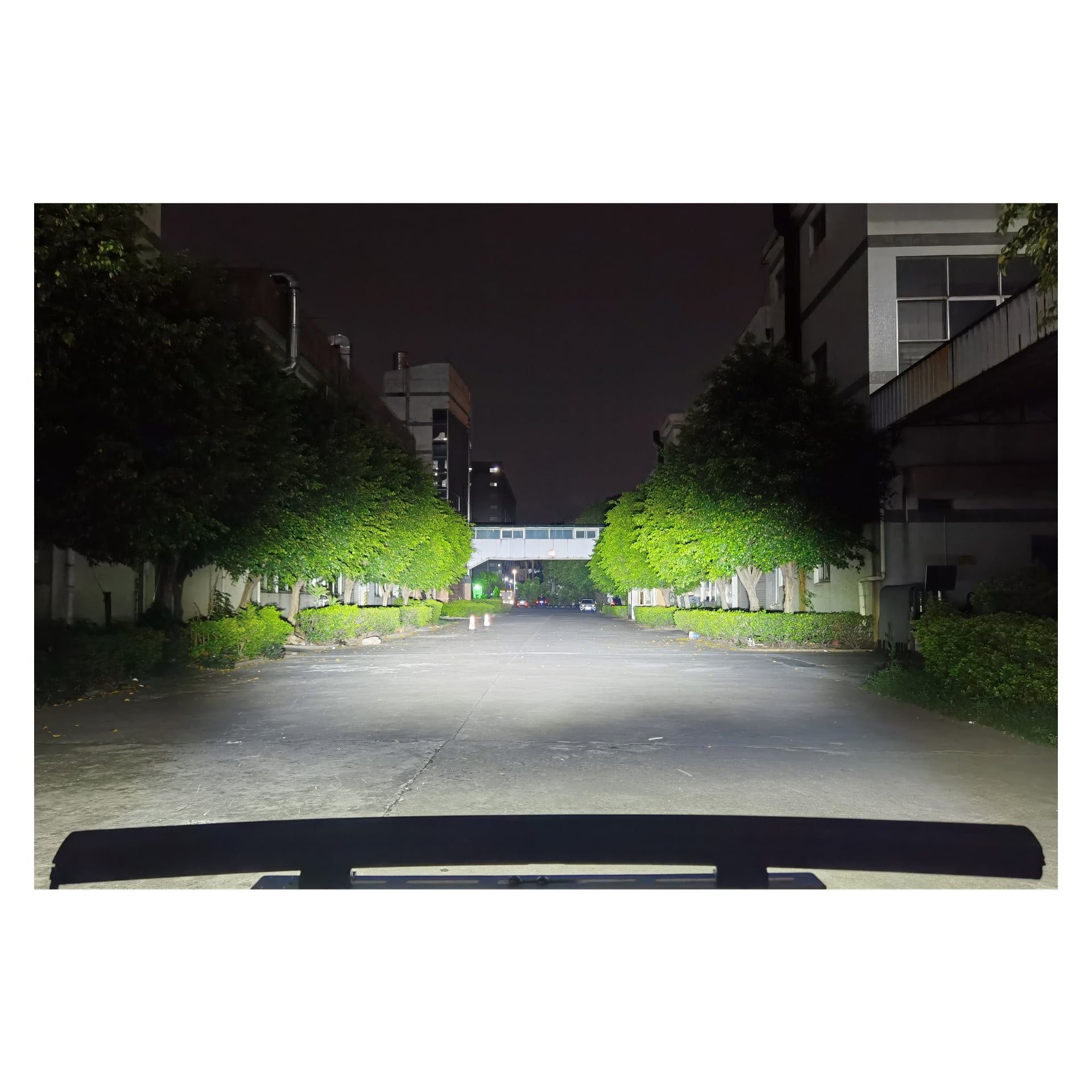 En tom, godt opplyst gate om natten har trær og bygninger på hver side. Hamron Light bar LED-frontlykter kaster sterkt lys over stedet, og en overbygd gangvei krysser to bygninger i det fjerne.
